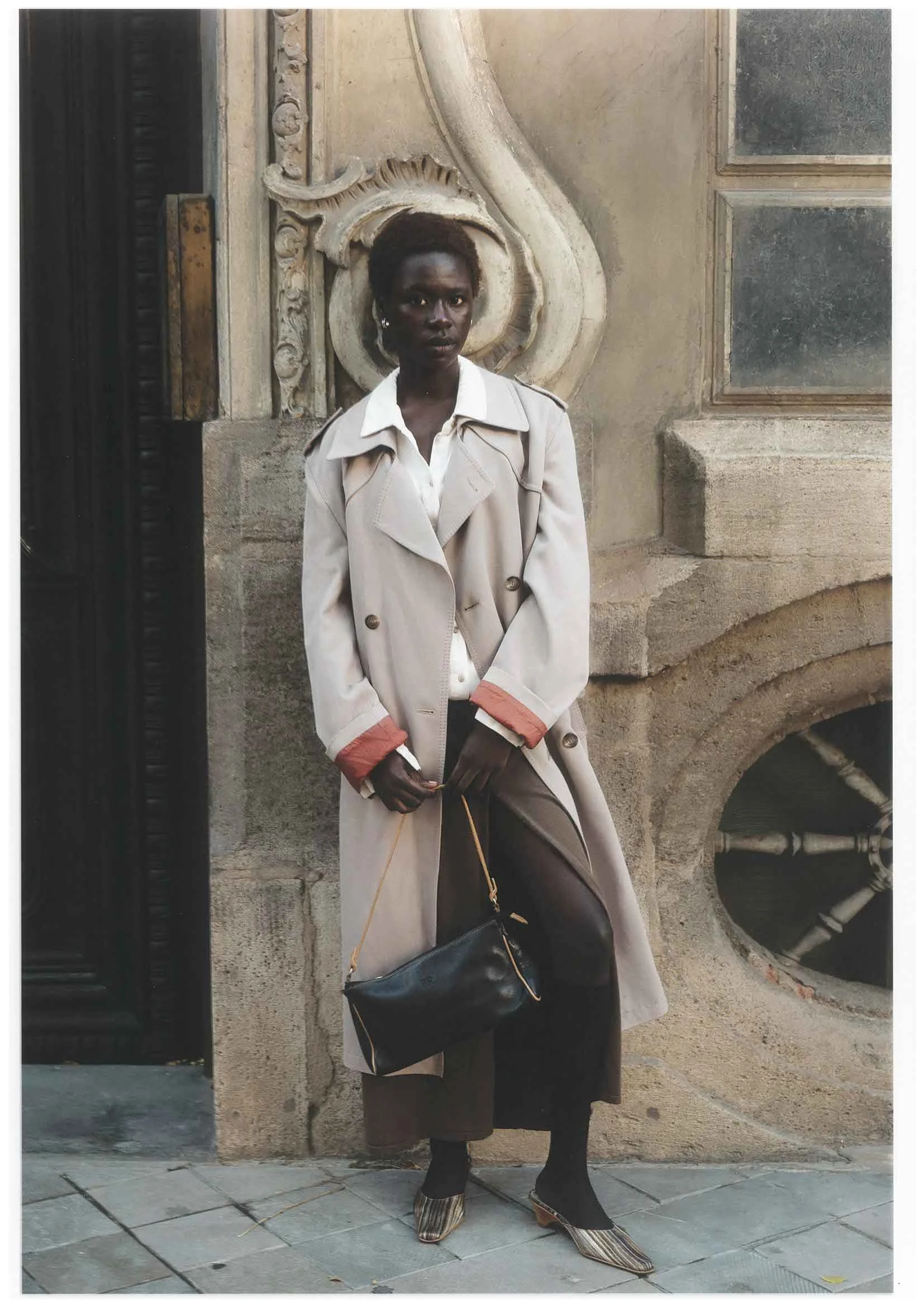 A woman standing in front of a historic building with ornate stone carving and small windows, wearing a beige trench coat, black leggings, patterned shoes, and holding a black handbag.