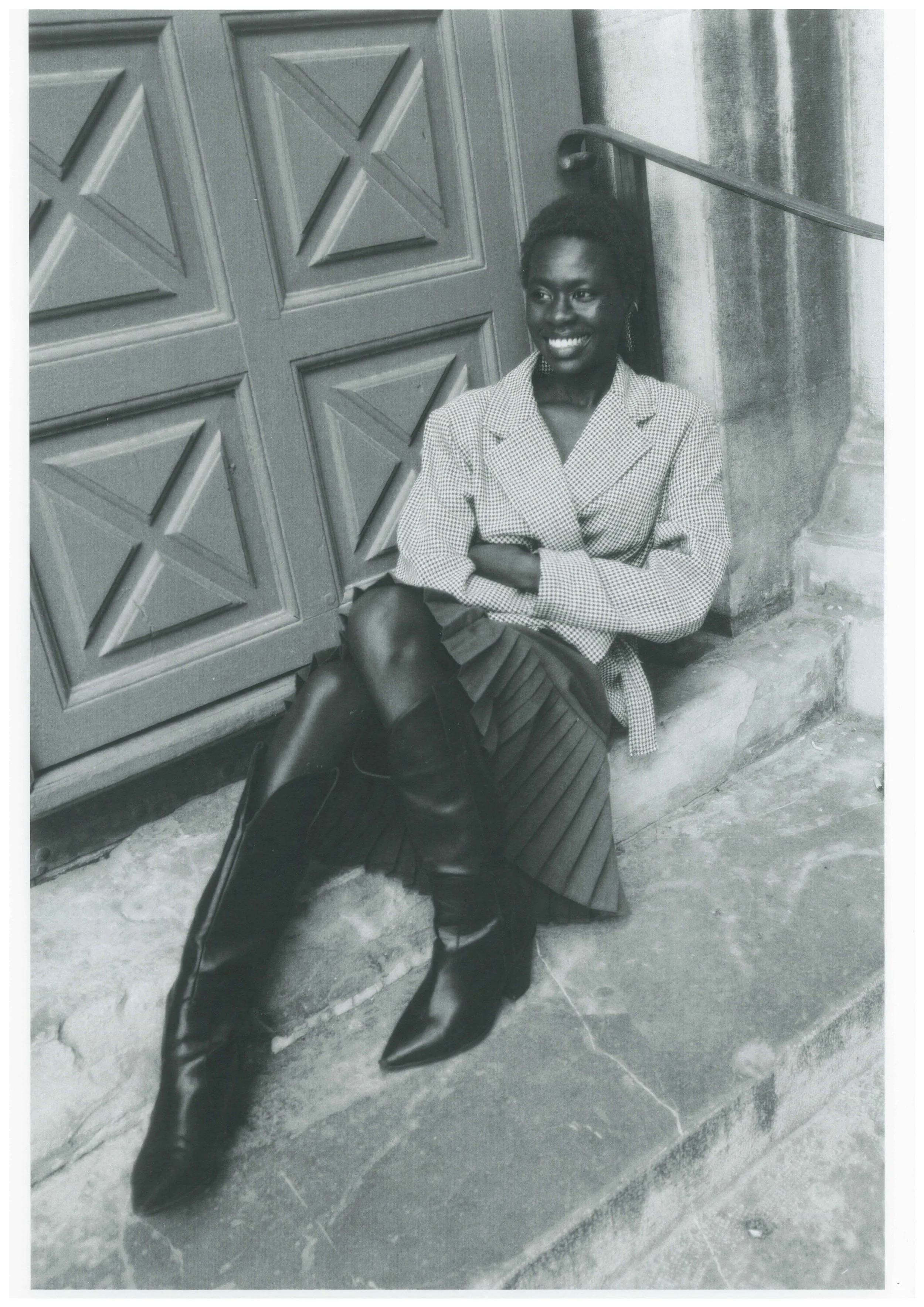 A woman sitting on a sidewalk leaning against a wall, smiling and looking to the side. She is wearing a patterned blazer, a pleated skirt, and knee-high boots.
