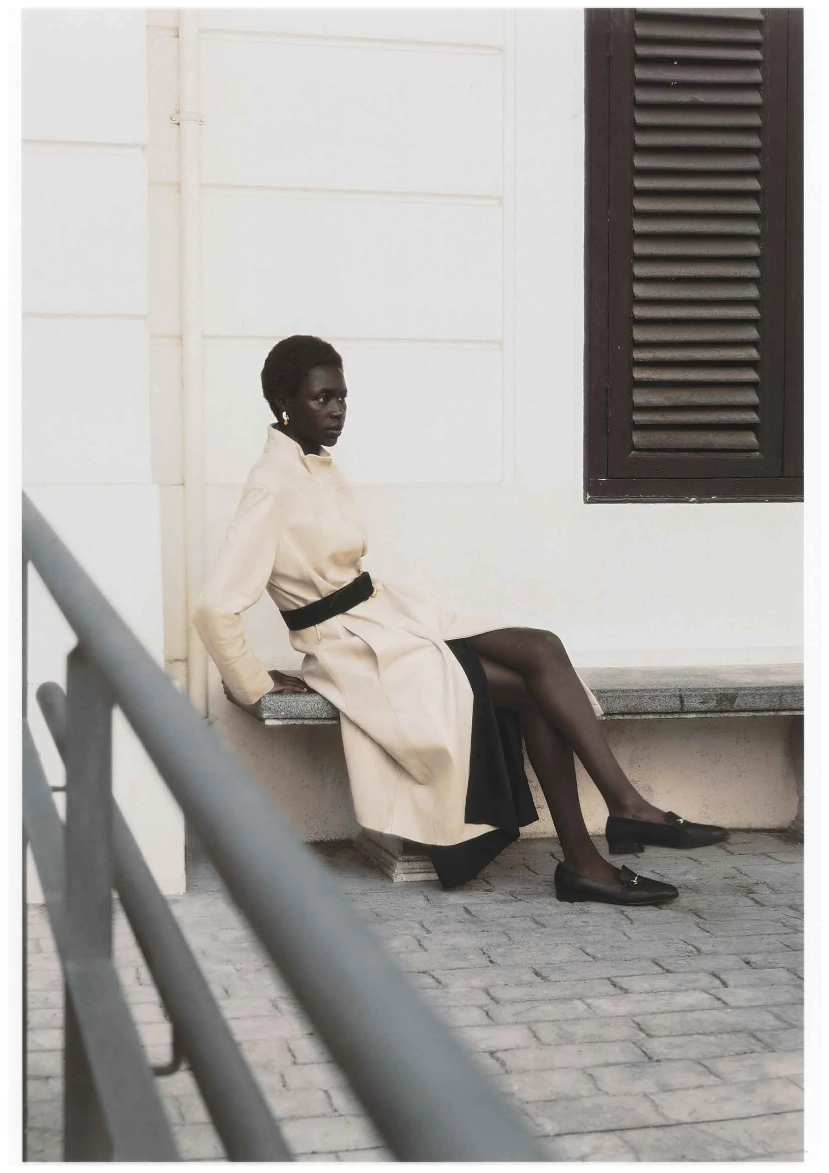 A woman sitting on a bench outside with a white wall behind her and a dark window to the side. She is dressed in a beige coat, black skirt, black tights, and black shoes, looking to the left.