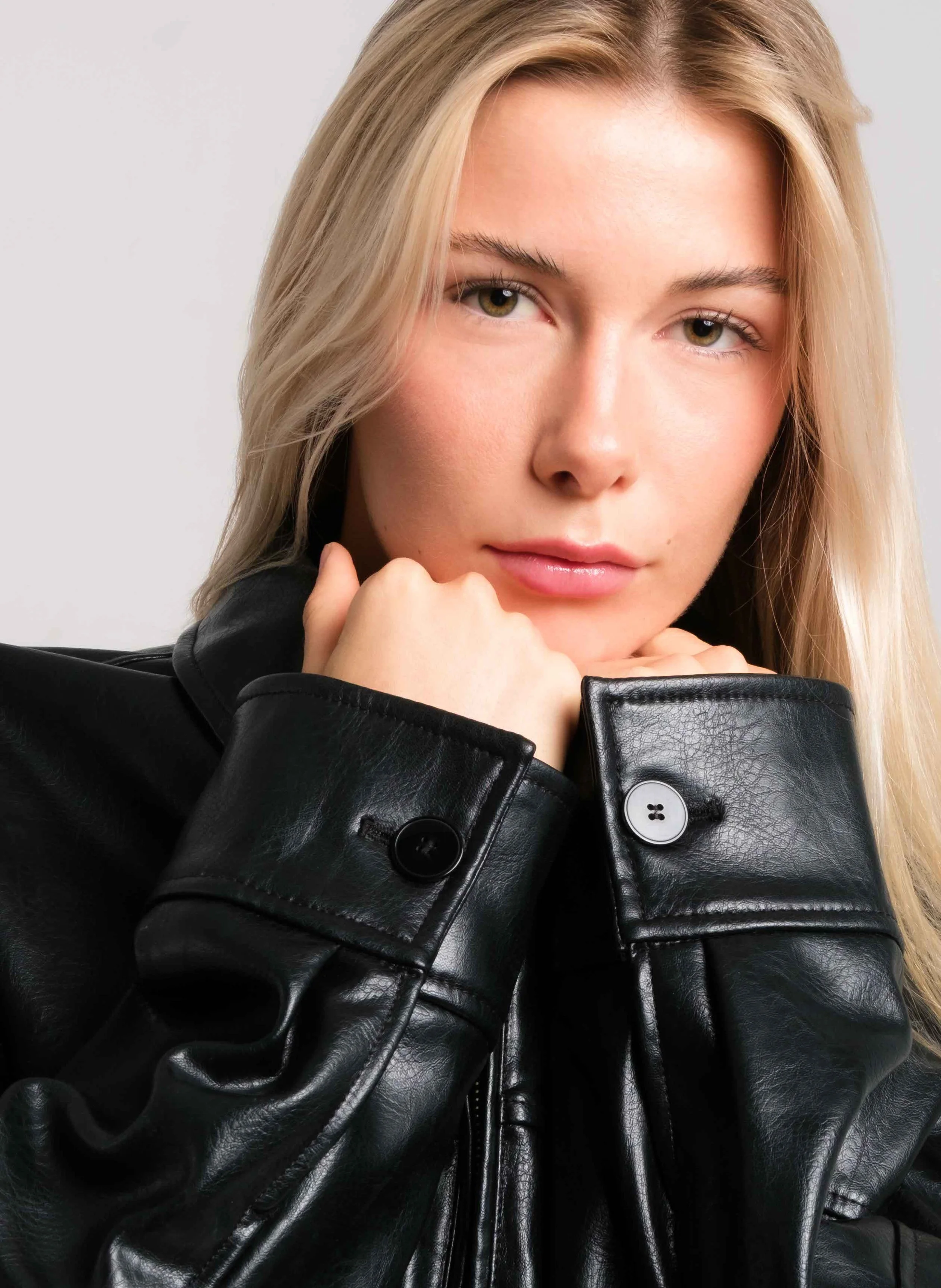 Close-up portrait of a young woman with blonde hair, wearing a black leather jacket, resting her chin on her hand, with a neutral facial expression.