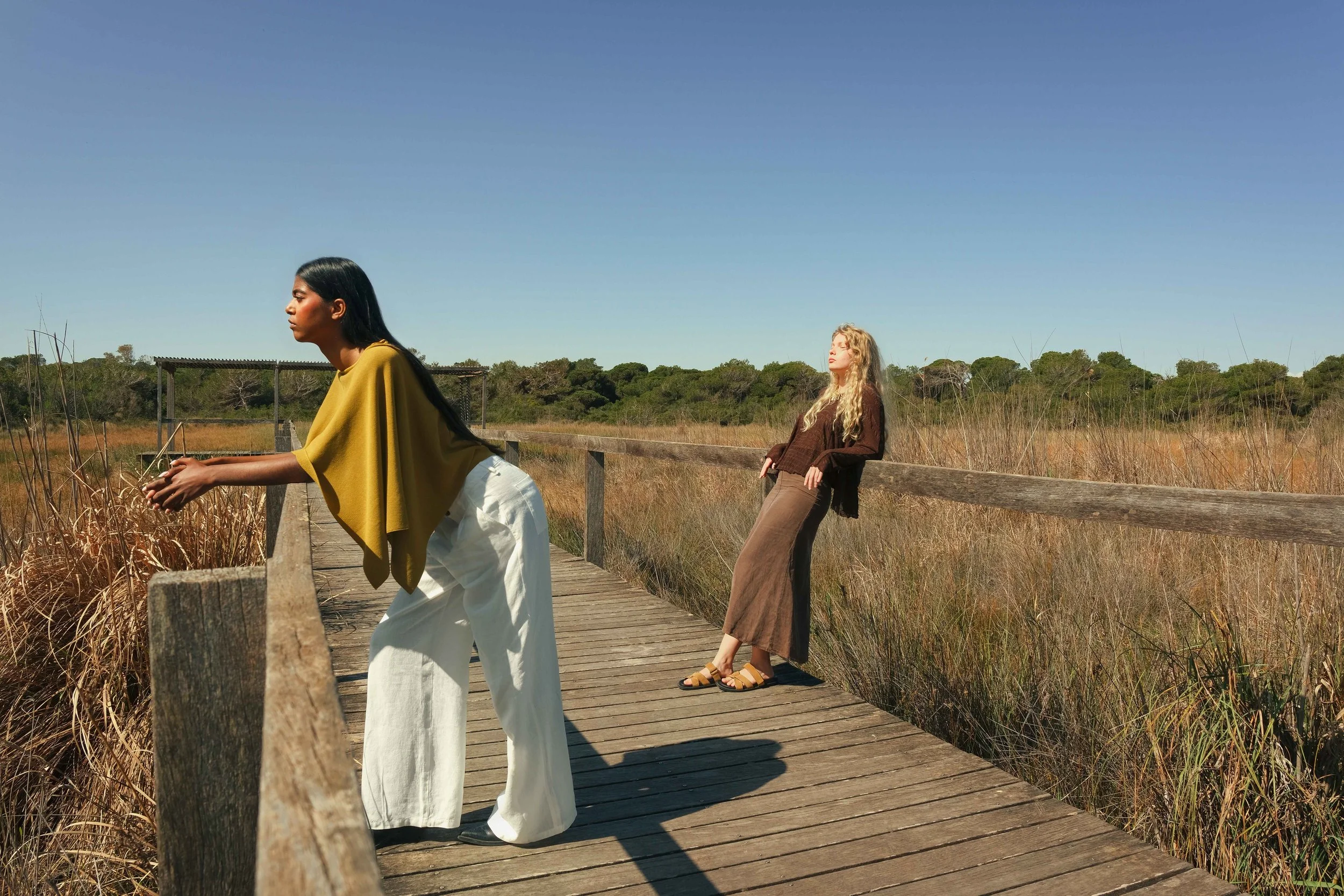Two women standing on a wooden bridge in a grassy field, with one leaning on the railing and the other leaning back against it, under a clear blue sky.