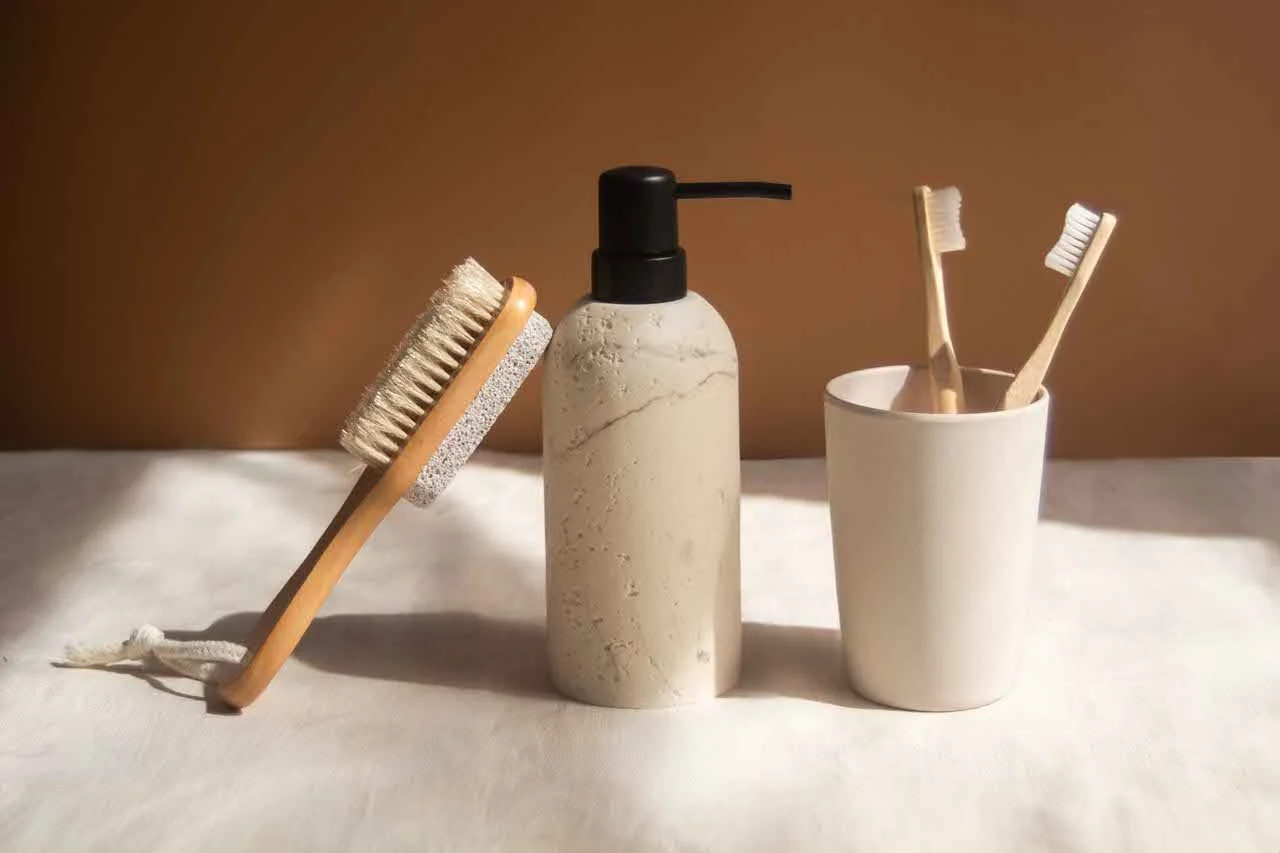Set of three bathroom items: a wooden-handled scrub brush, a marble-patterned soap or lotion dispenser, and two toothbrushes in a white cup.