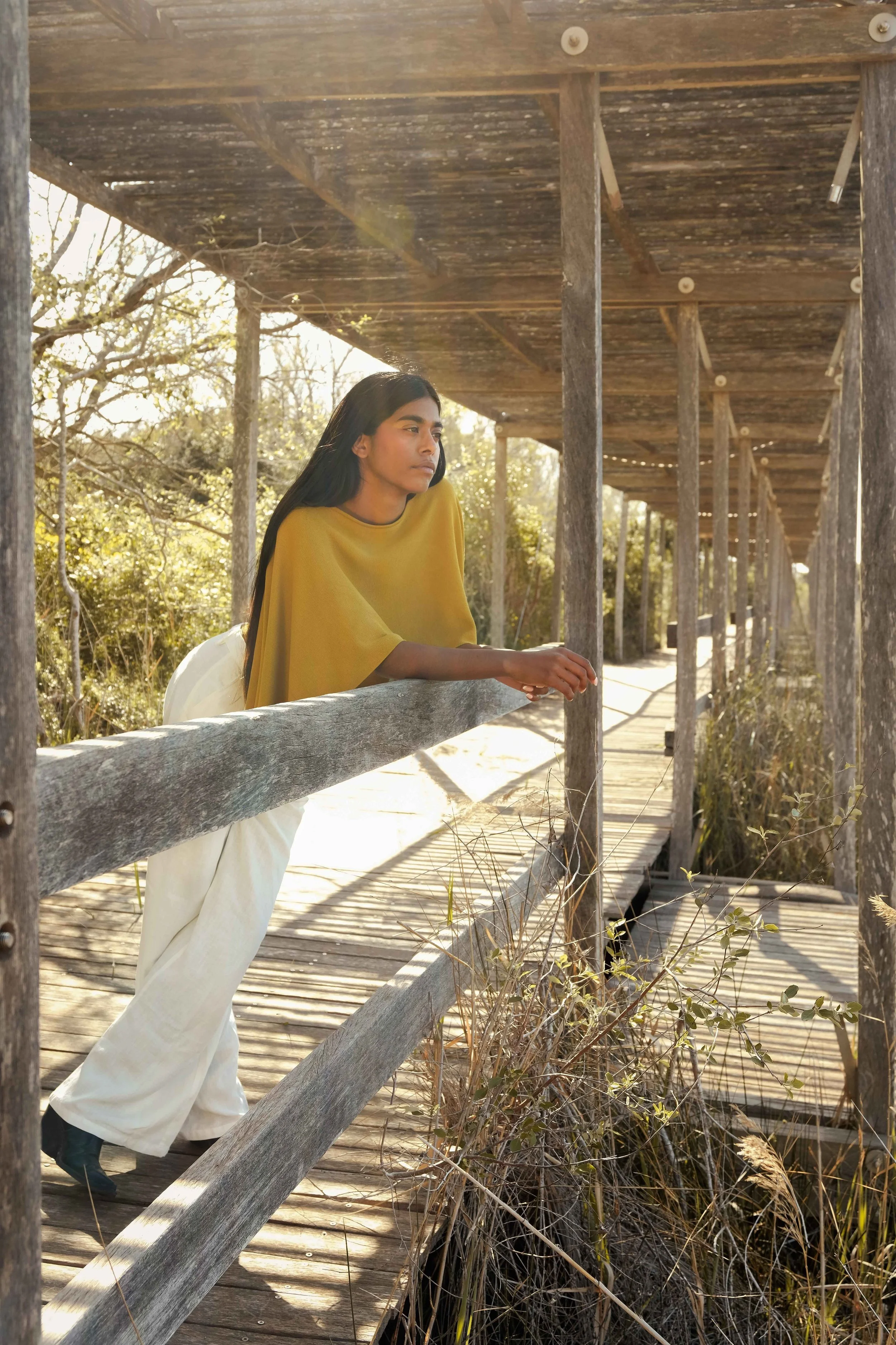 A woman with long black hair leaning on a wooden railing on a weathered wooden boardwalk, wearing a mustard yellow top and white pants, looking thoughtfully into the distance with sunlight illuminating her face and surroundings.
