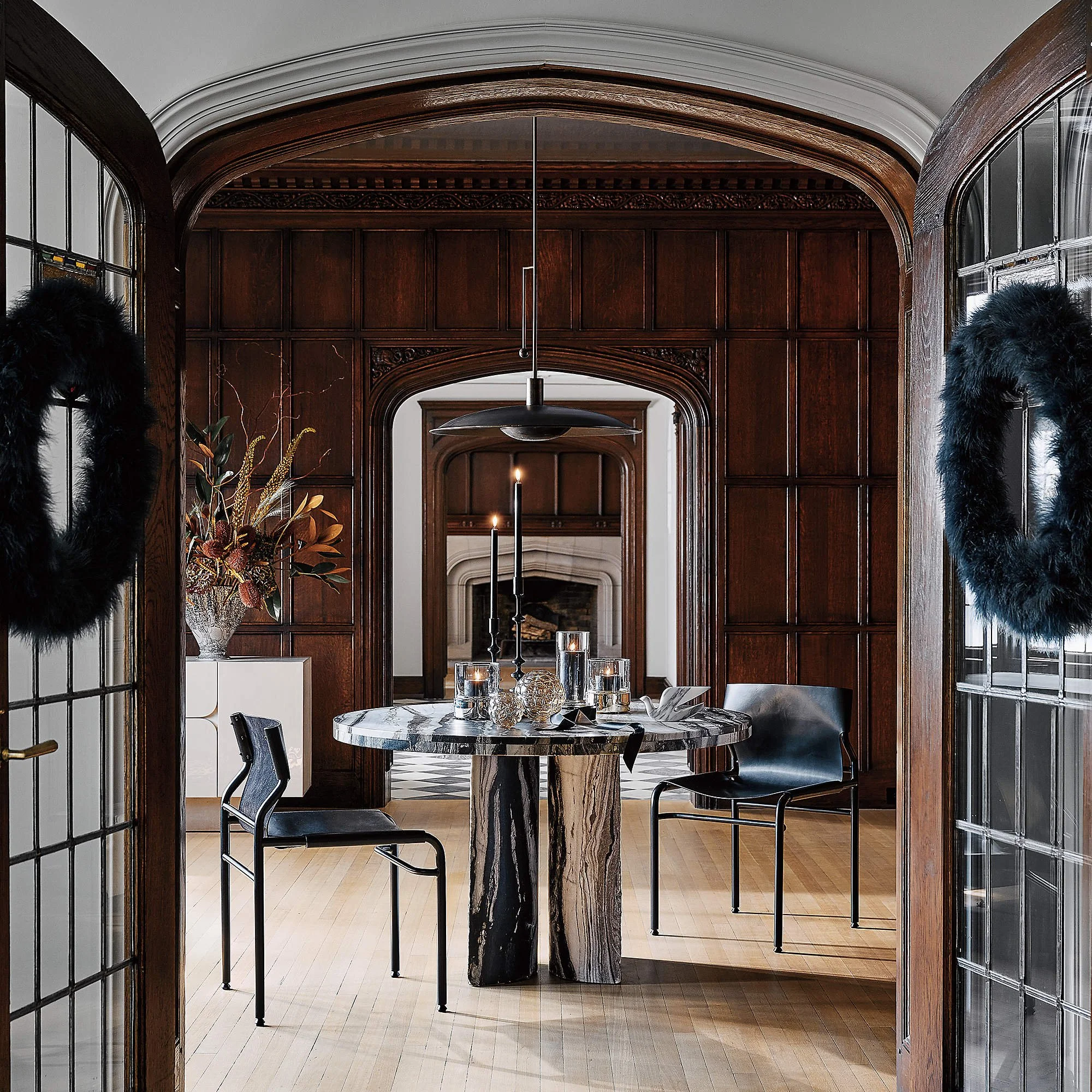 A stylish dining room with a marble table, black chairs, candles, and decorative objects, framed by wooden archways with black wreaths on glass doors.