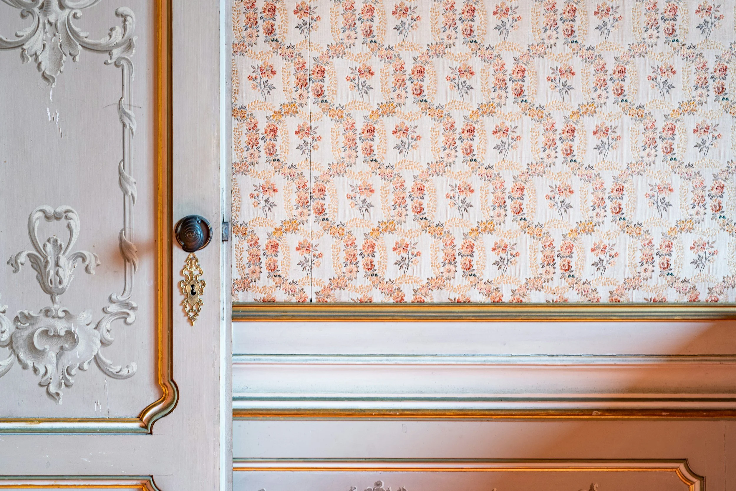 Interior of a room with a floral-patterned wallpaper in shades of orange, beige, and gray. The wall features detailed white decorative molding and trim painted in a gold hue.