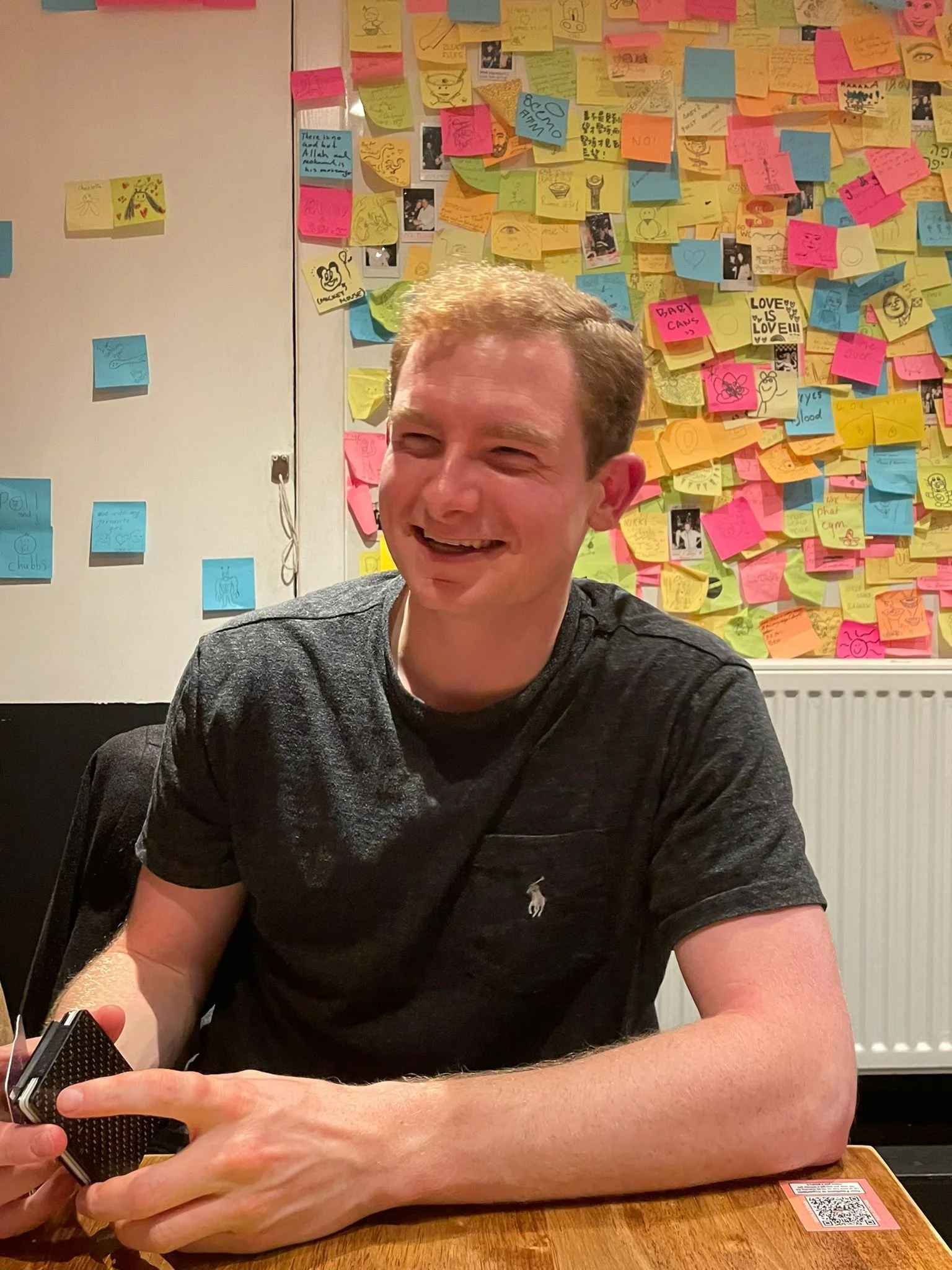 A smiling man in a black shirt sitting at a wooden table, holding a small black object with a textured surface in his hands, with a colorful wall of sticky notes and photographs in the background.