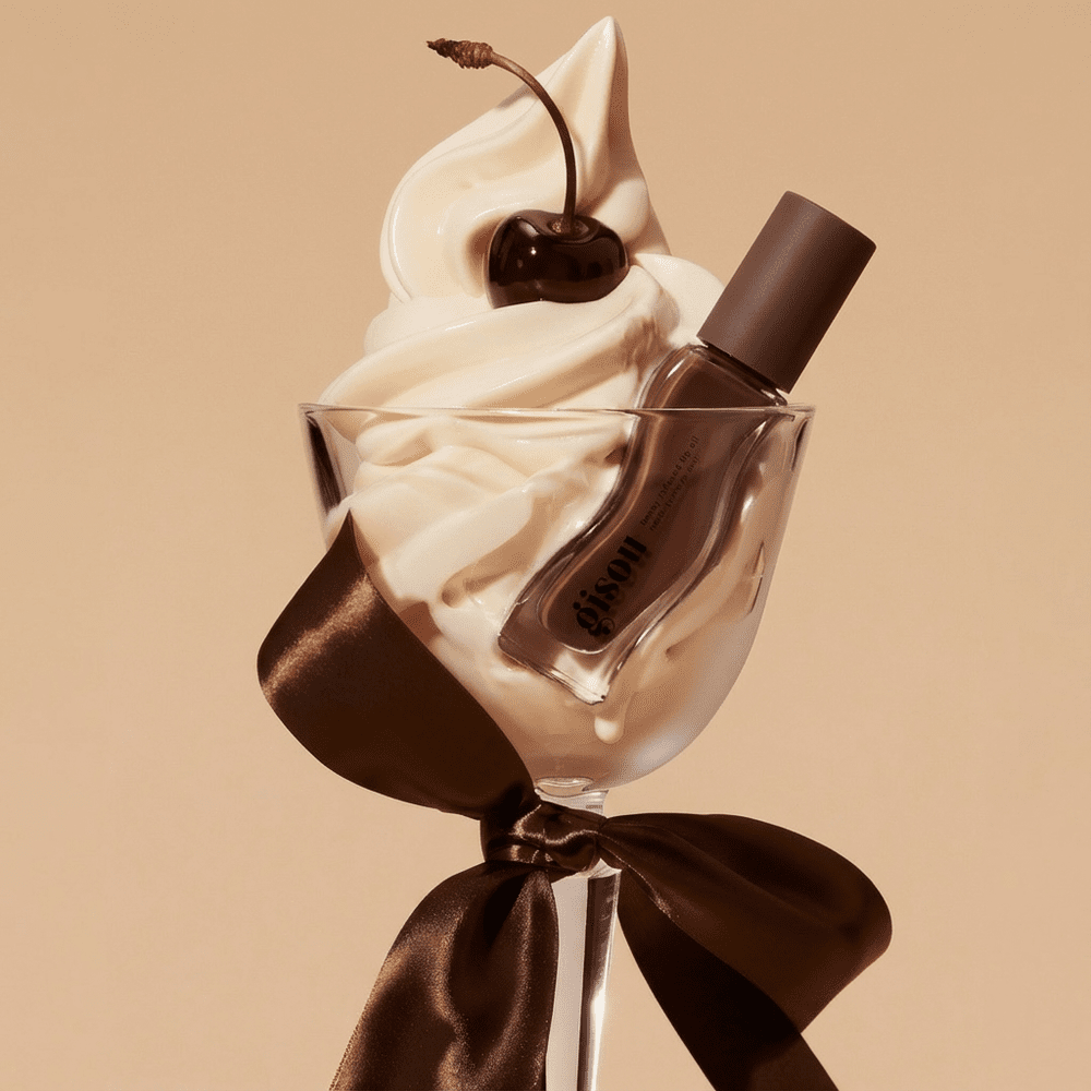 Een glazen dessertcoupe gevuld met slagroom, afgewerkt met een kers en versierd met een Gisou-lipgloss, alles samengebonden met een bruin satijnen lint.