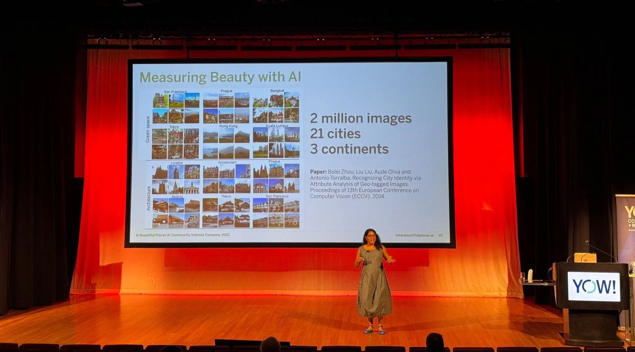 Chanuki Seresinhe giving a presentation on stage at a conference. Slide titled 'Measuring Beauty with AI,' featuring sample images of cityscapes from multiple cities around the world, and text mentioning 2 million images, 21 cities, and 3 continents.