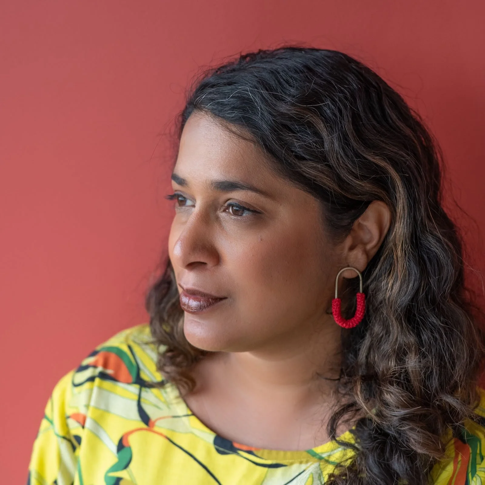 Close-up of Chanuki Seresinhe wearing a yellow floral top and red beaded hoop earrings, looking to the side against a coral background.
