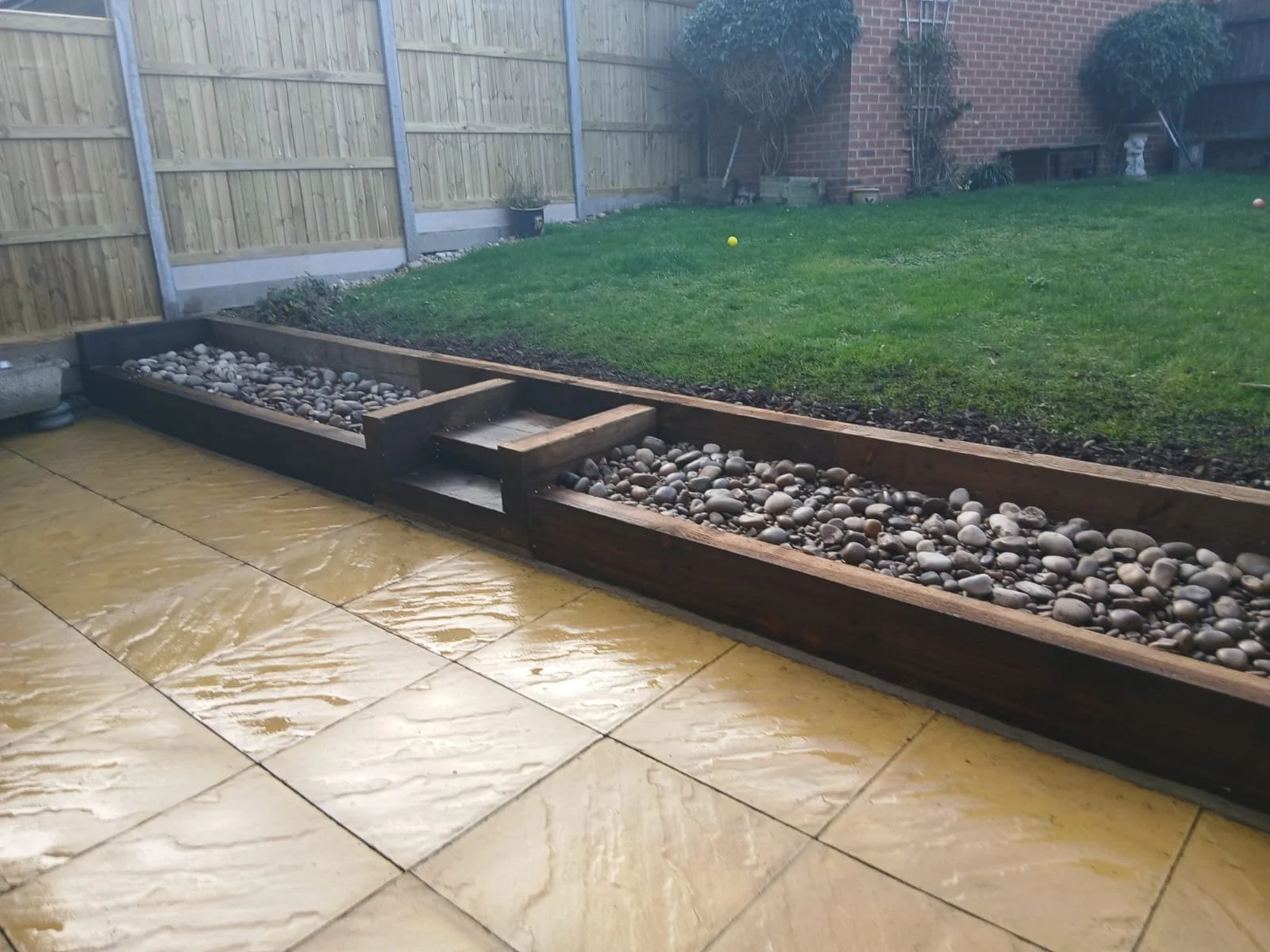 View of a backyard with a paved patio, a grass lawn, a wooden fence, and a brick house wall. There are raised garden beds with rocks for drainage, and a small staircase in one of them. Several tennis balls are scattered on the grass.