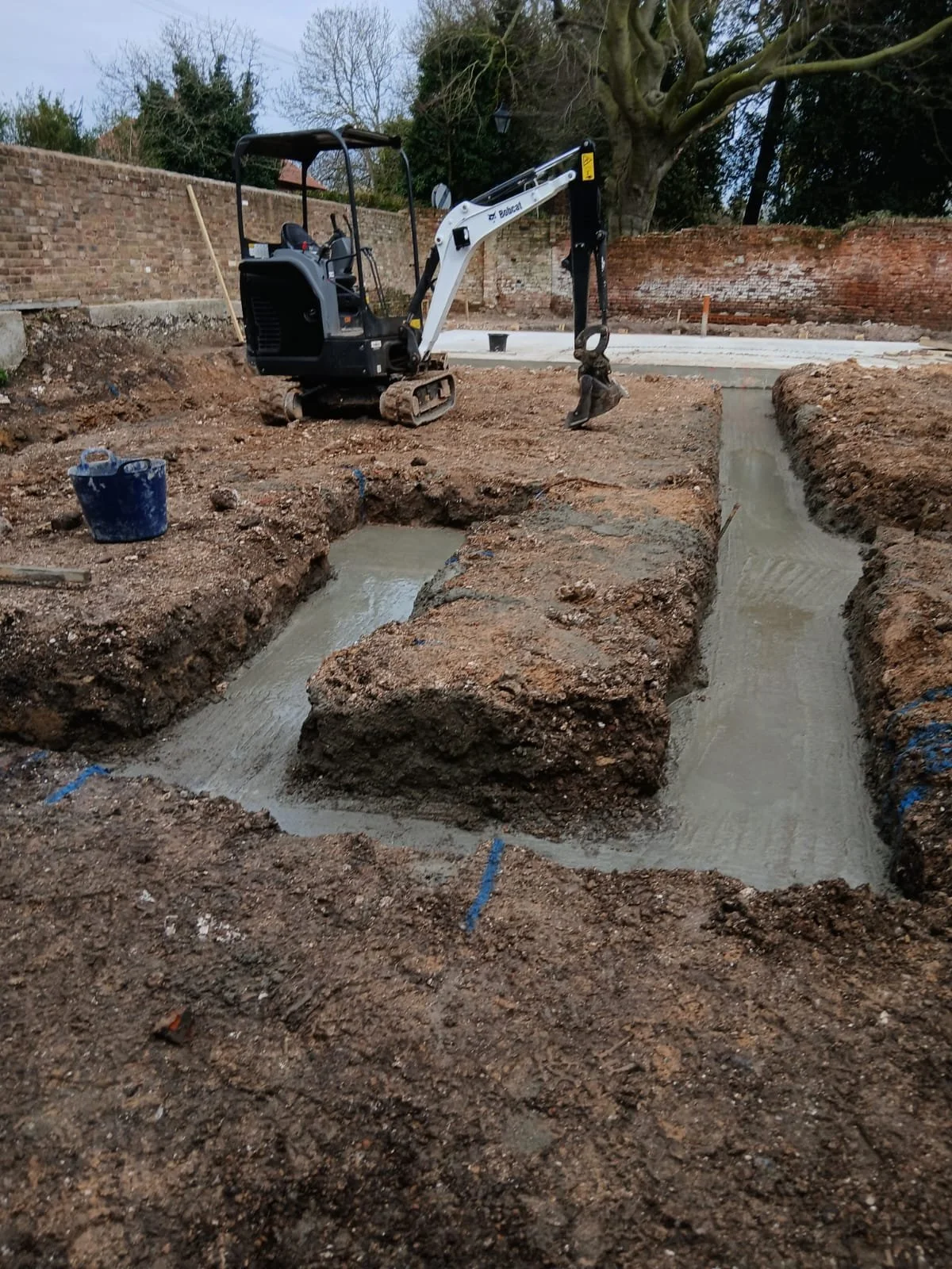 Construction site with excavated trenches and a mini excavator. The trenches are lined with wet concrete, and there are blue markings on the ground. In the background, there are brick walls and trees.