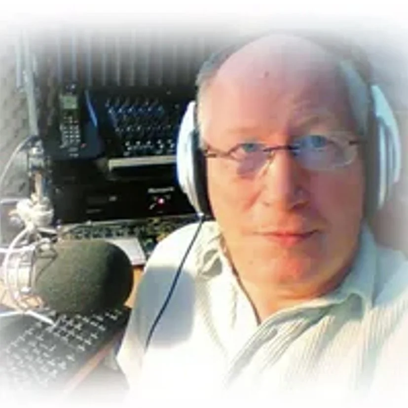 A man wearing glasses and headphones sitting in front of radio broadcasting equipment, including a microphone, sound mixer, and other electronic devices.
