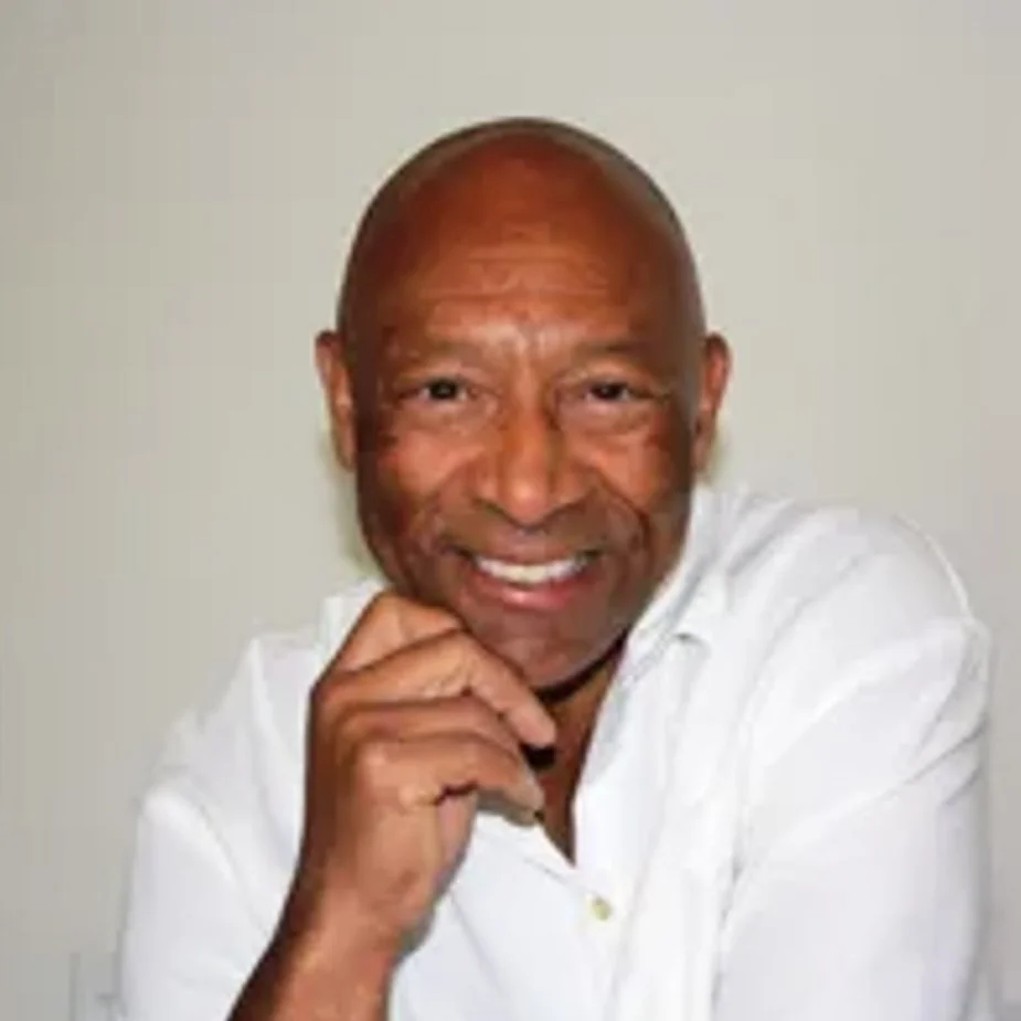 A smiling bald man in a white shirt, posing with his hand near his chin against a plain background.