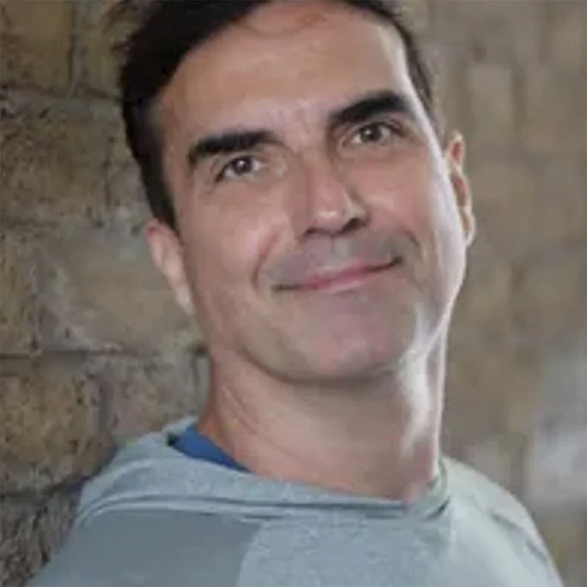 Close-up of a smiling man with dark hair, wearing a gray shirt, standing against a brick wall background.