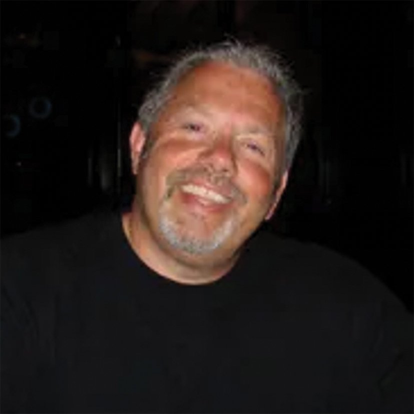A smiling man with gray hair and a beard, wearing a black shirt, in a dark setting.