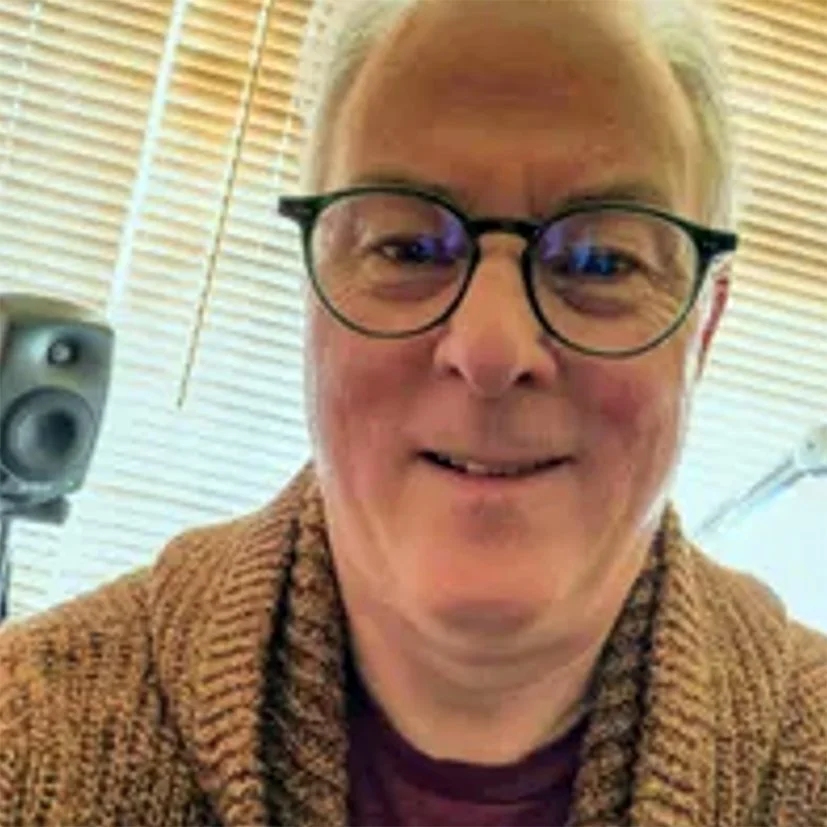 A smiling middle-aged man with glasses and short, light-colored hair, wearing a knitted brown sweater, sitting indoors near wooden blinds with a speaker in the background.