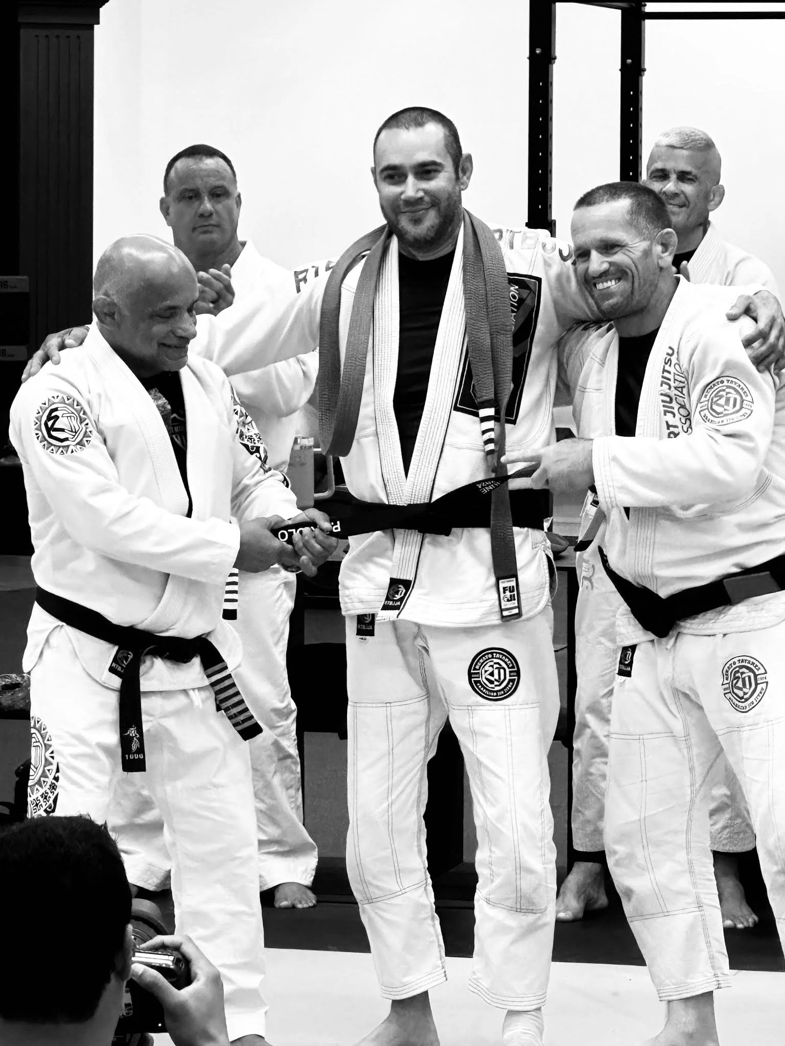 Group of five black and white photo of Brazilian Jiu-Jitsu practitioners in gi, with one receiving a black belt, in a dojo.