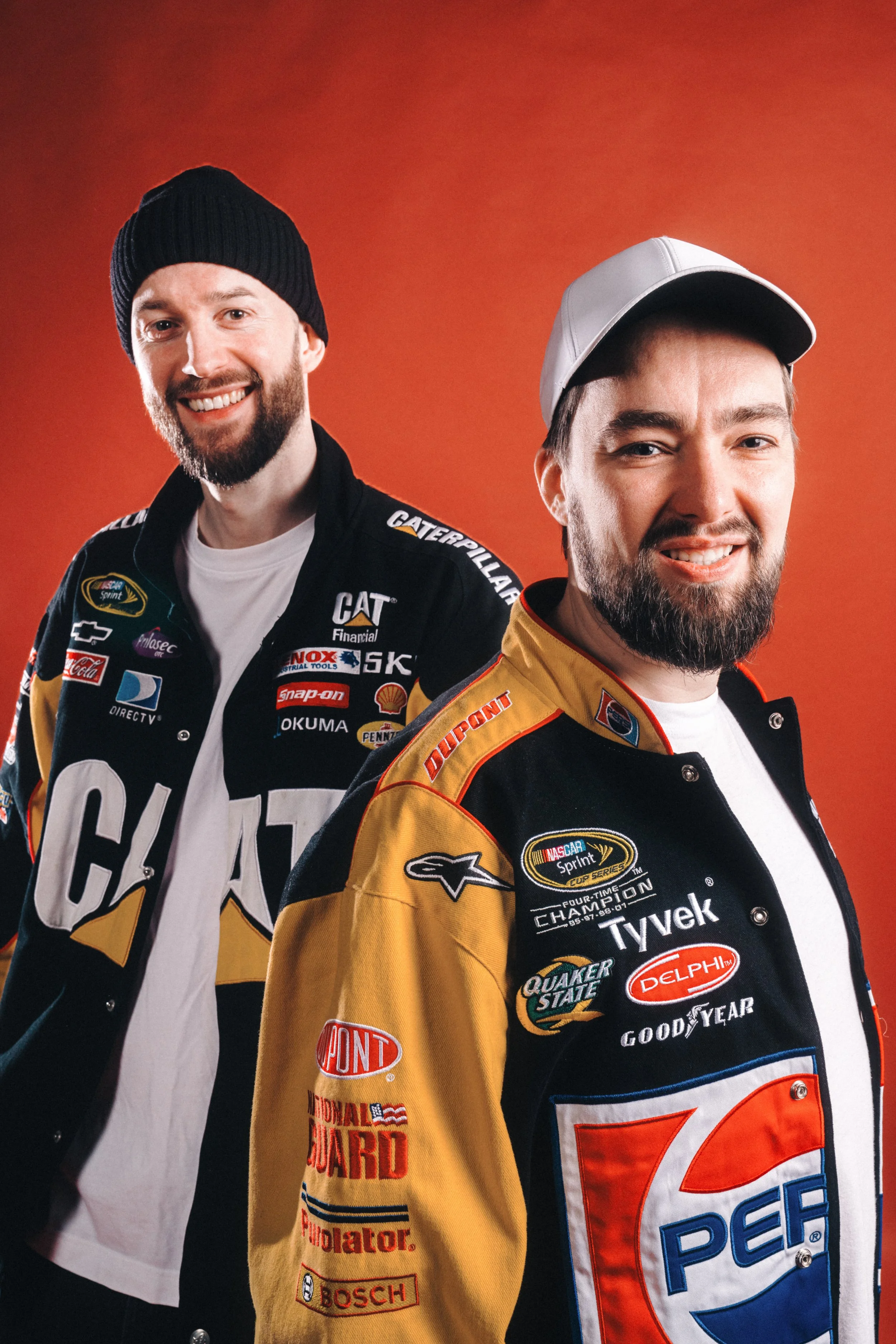 Two men wearing racing jackets with sponsor logos, smiling against a red background.