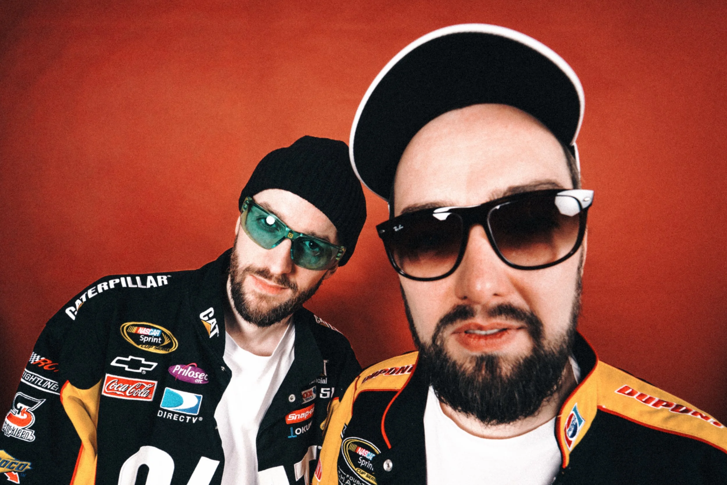Two men in racing jackets, sunglasses, and hats or beanies, standing in front of a red background.