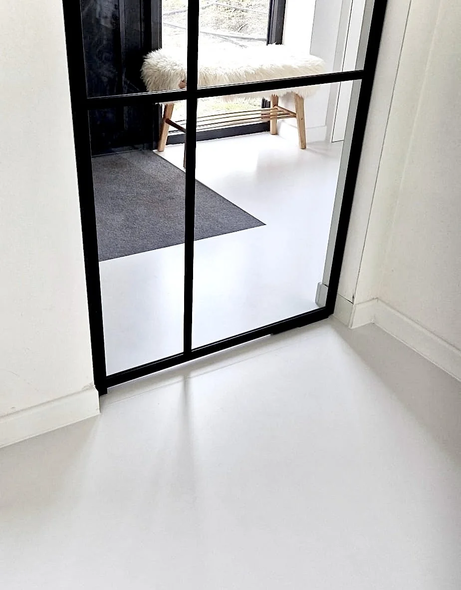 View through a black-framed glass door into a room with white flooring, a dark gray rug, a bench with a fluffy white seat, and a sliding glass door leading outside.