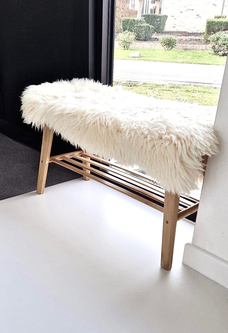 A wooden bench with a fluffy white sheepskin cover, placed near a glass door with a view of a garden with bushes and grass.