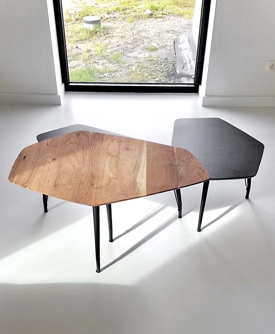 A modern interior with a white floor and minimal decor, featuring a unique two-tone coffee table with one wooden and one fabric surface, and black metal legs, positioned near a large glass door leading outside.