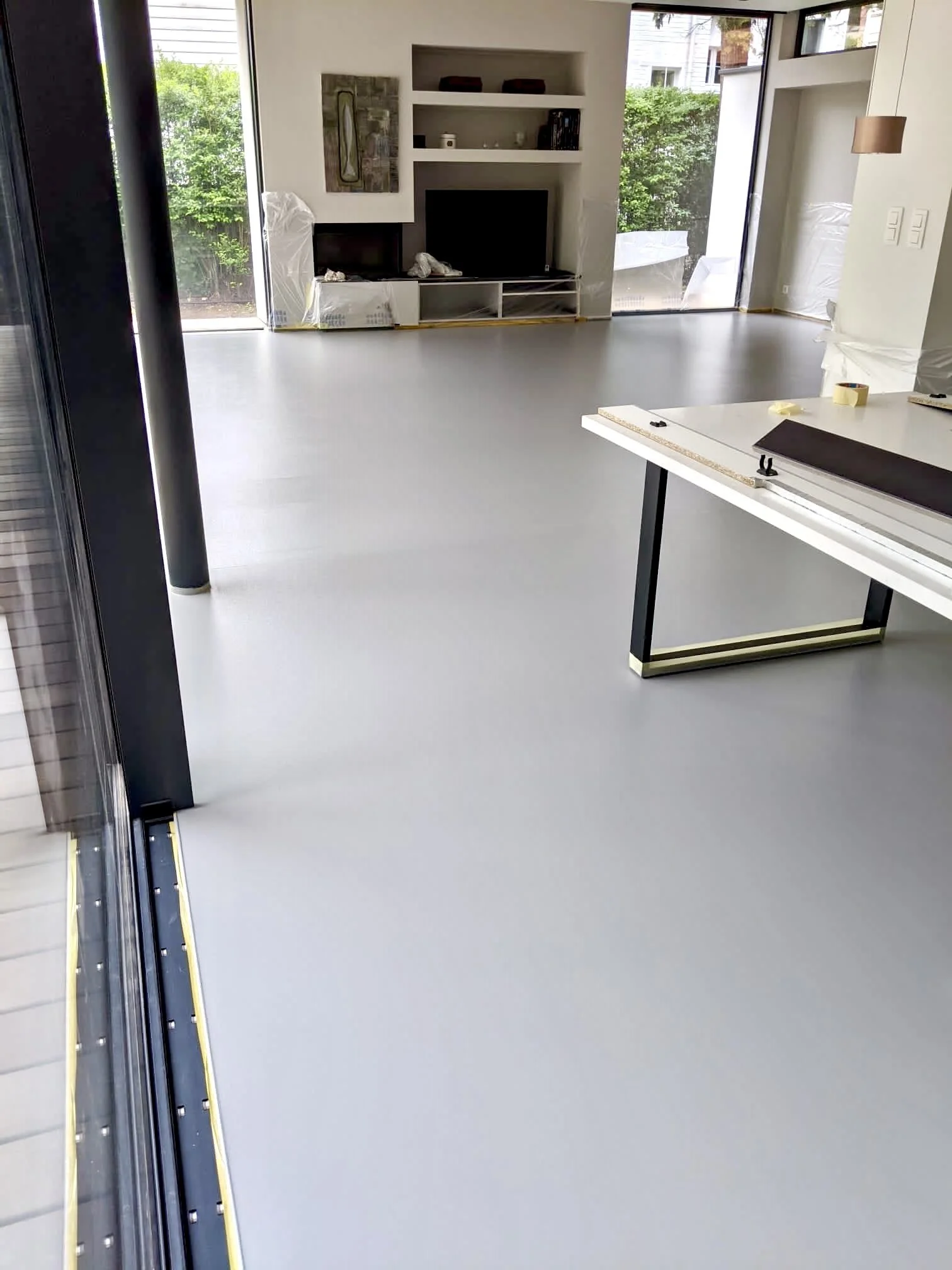 Modern living room with large windows, built-in shelves, wall-mounted TV, and white flooring under construction or renovation.