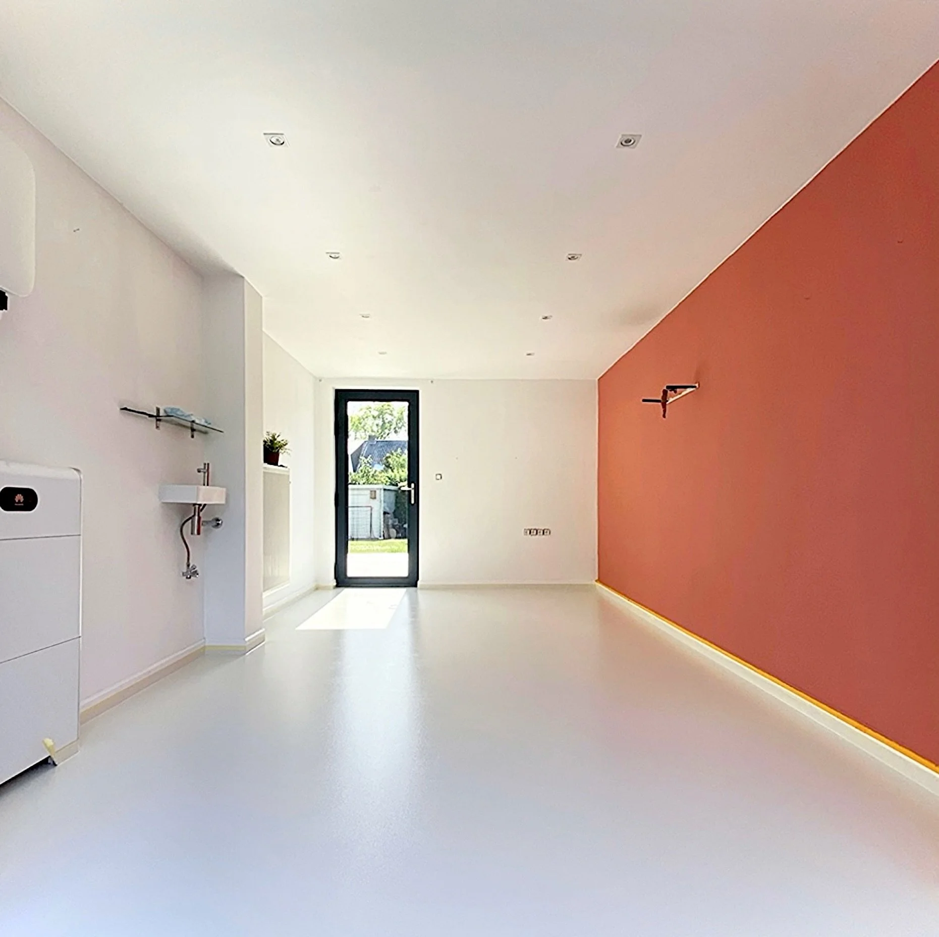 Empty interior room with white flooring, white ceiling, and a single orange accent wall. A black framed glass door leads outside, and there is a small sink on the left wall along with a wall-mounted shelf. The room appears to be under construction or