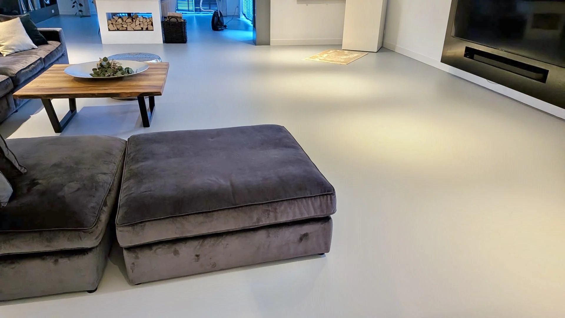 Living room with gray couch and ottoman, wooden coffee table with decorative centerpiece, fireplace with logs, and a modern, minimalistic interior design.