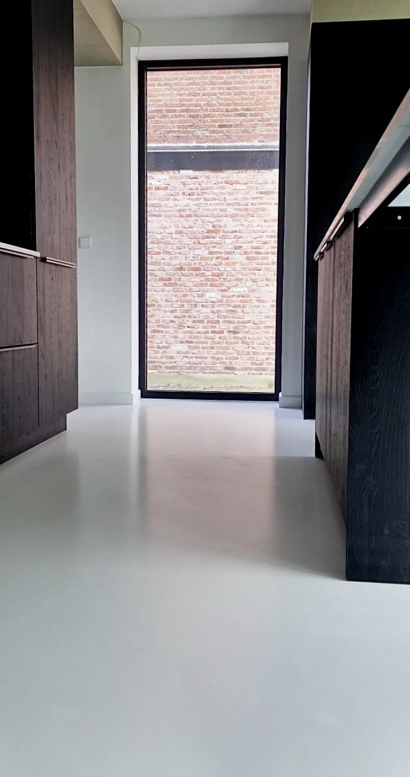 View of a modern kitchen with dark wood cabinets, white flooring, and a tall glass door leading outside to a brick wall.