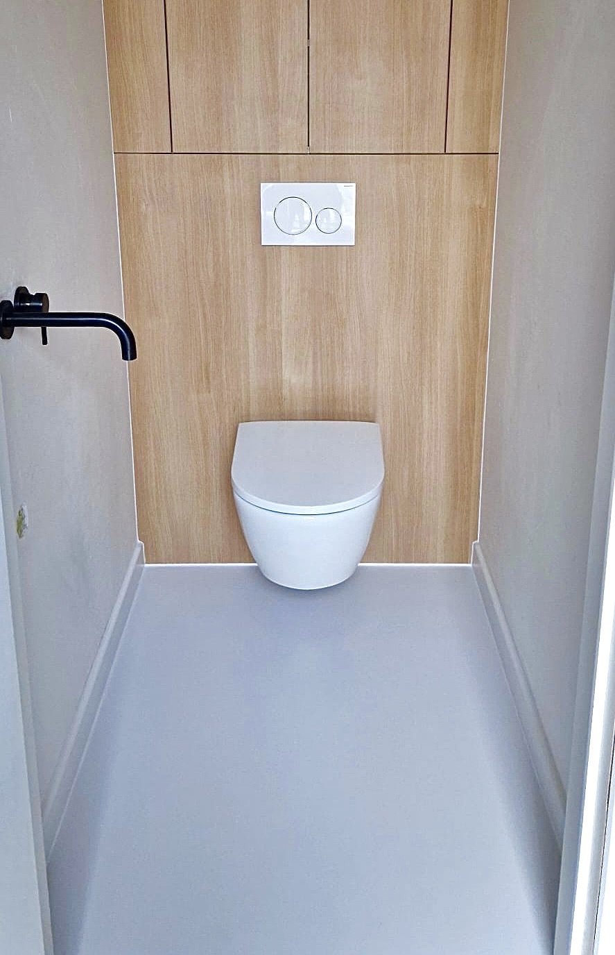 A modern toilet with a concealed tank, mounted on a wooden-paneled wall, in a minimalistic bathroom with a white floor and black faucet on the left side.