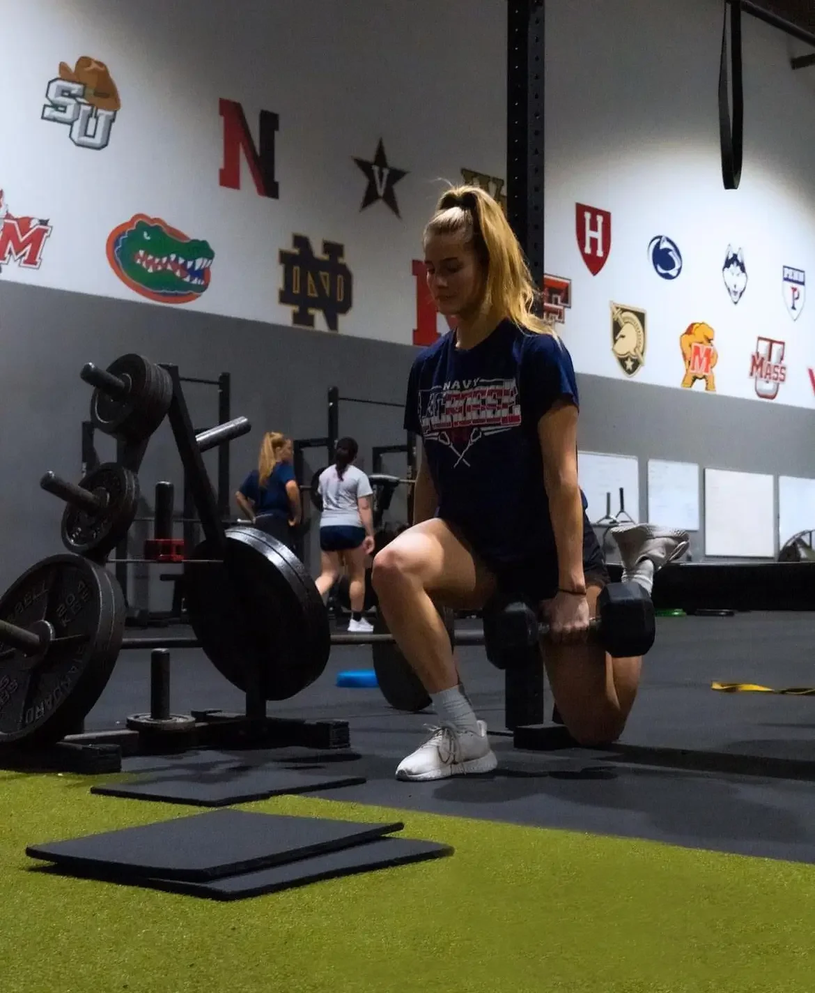 A woman in a gym performing a lunge while holding dumbbells. She is wearing a blue T-shirt, white sneakers, and gray socks. In the background, other people are working out, and various sports team logos and mascots are displayed on the wall.