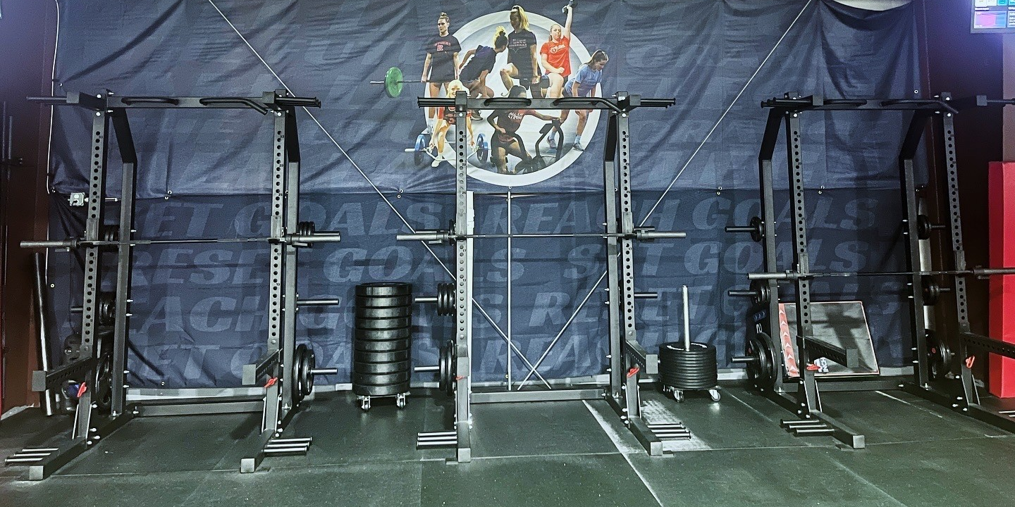 Empty gym area with three power rack stations, weight plates, and a black background with a fitness-themed mural.