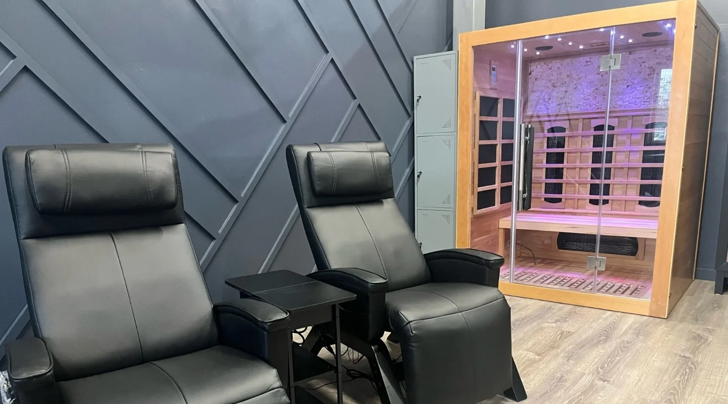 Two black massage chairs and a small black table between them, next to a wooden infared sauna with glass door and purple lighting inside, in a modern room with blue geometric wall panels and wood-like flooring.