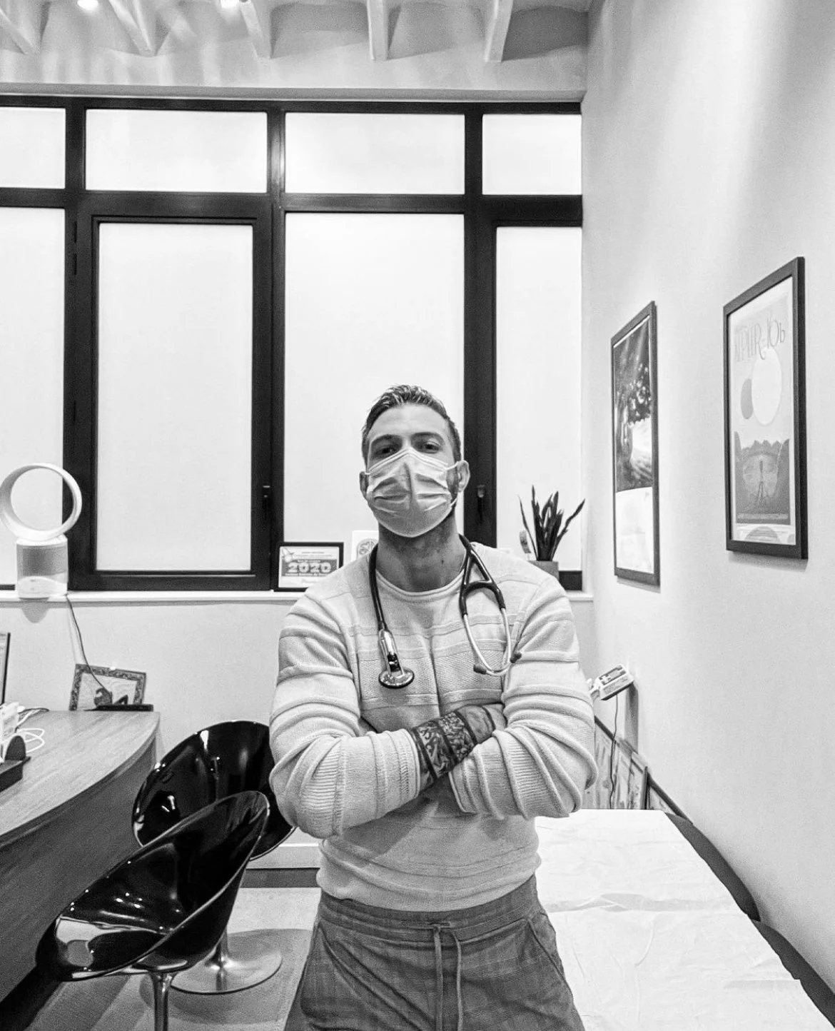 A man with a stethoscope around his neck wearing a face mask standing in a room with large windows, a desk, a chair, and framed posters on the wall. Médecin généraliste Français à Dubaï / GP / French GP / French Doctor / French family medicine