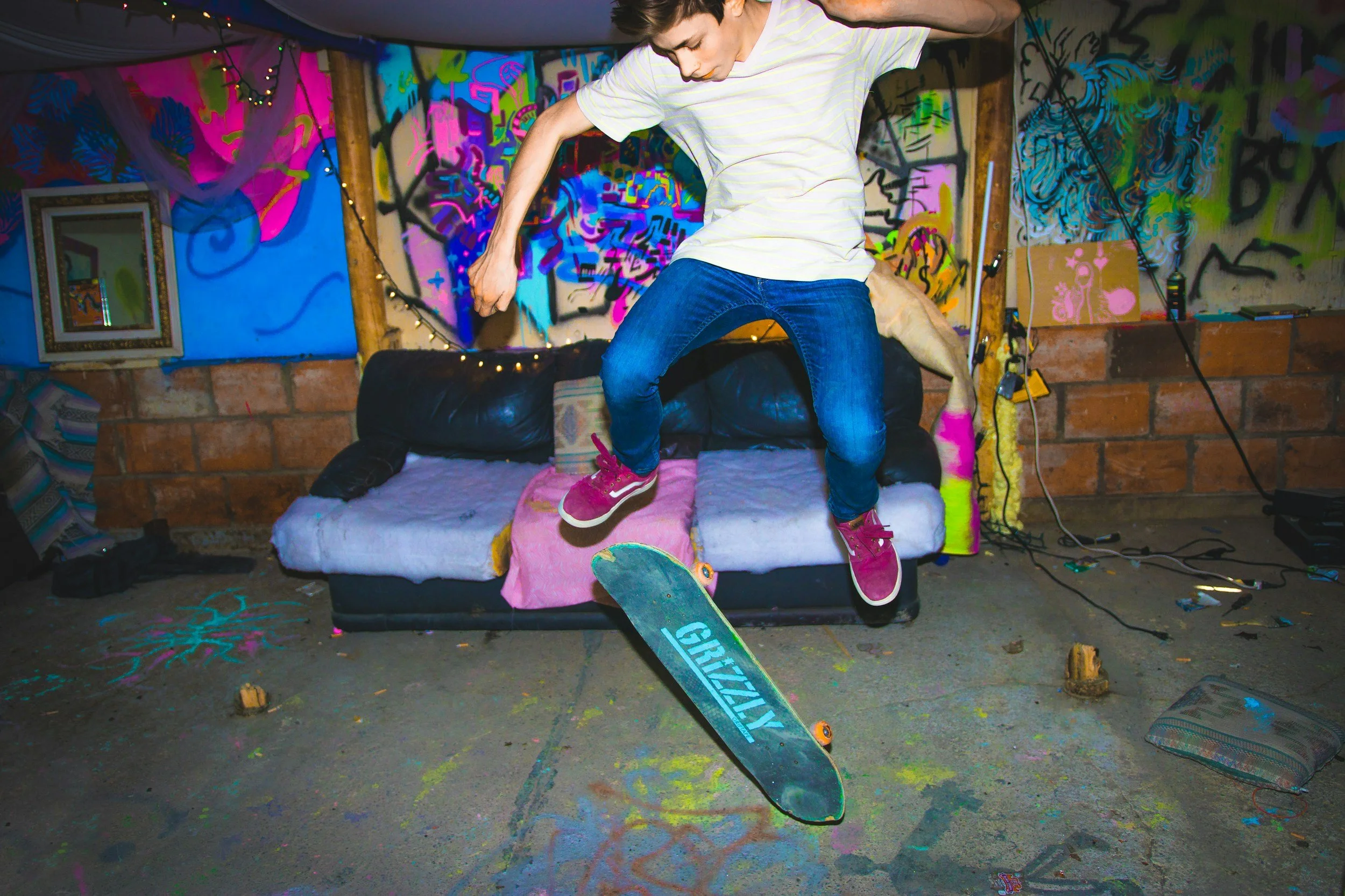 Person skateboarding indoors in a room with colorful graffiti art, a black couch, and various objects on the floor.