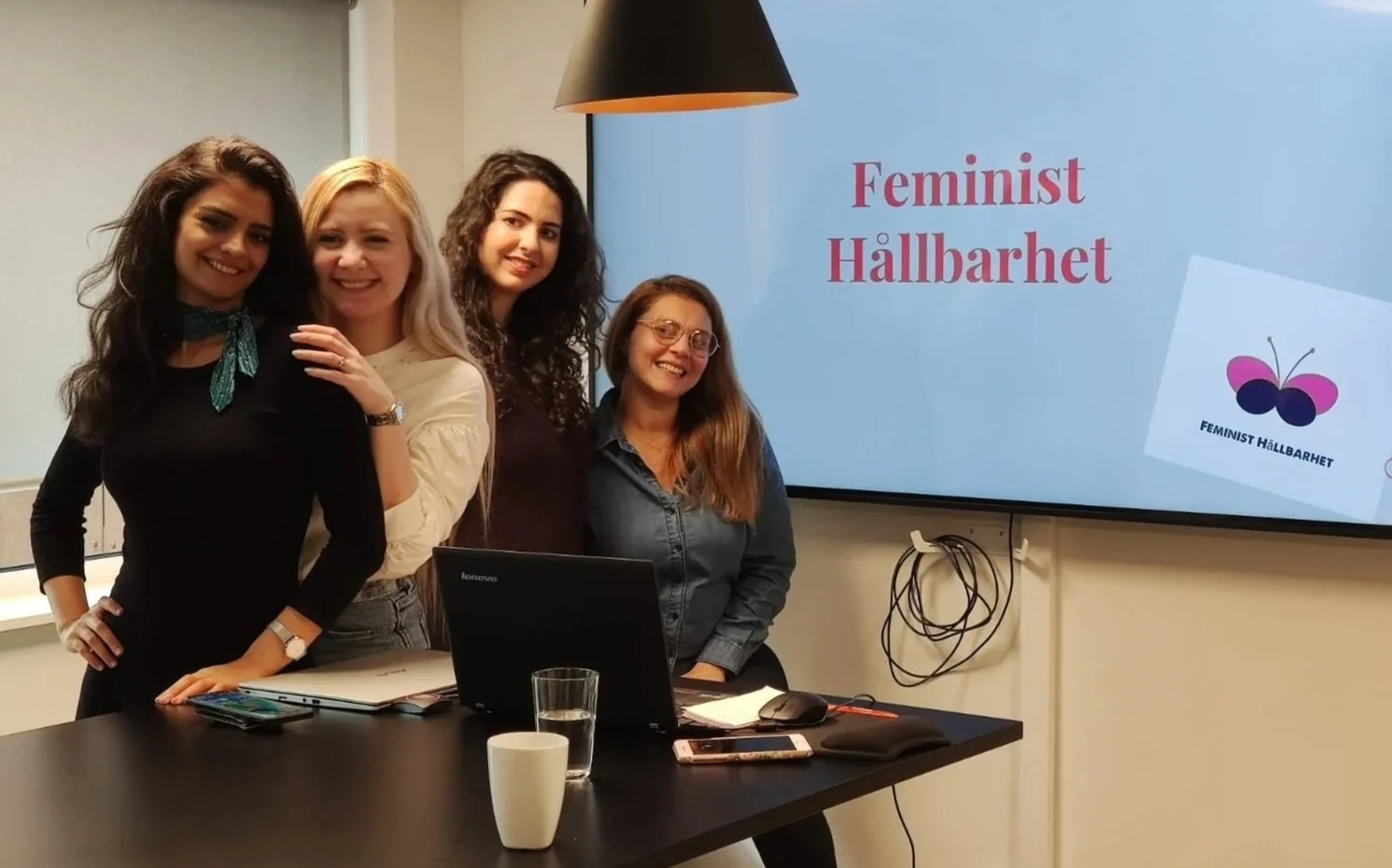 Four women standing together in a conference room, smiling, with a presentation screen behind them displaying 'Feminist Hållbarhet' and a butterfly logo.