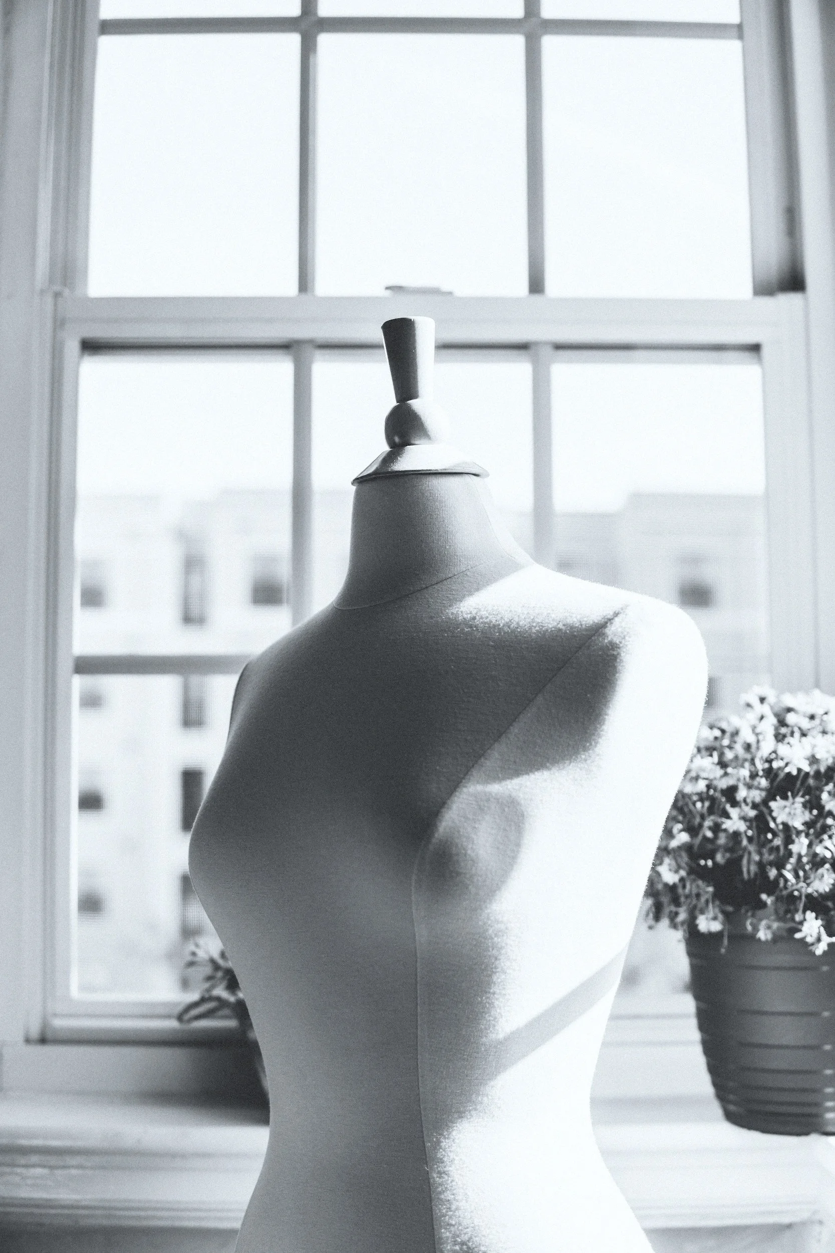 A dress form mannequin stands in front of a window with a cityscape view, next to a potted flowering plant, with natural light streaming in.