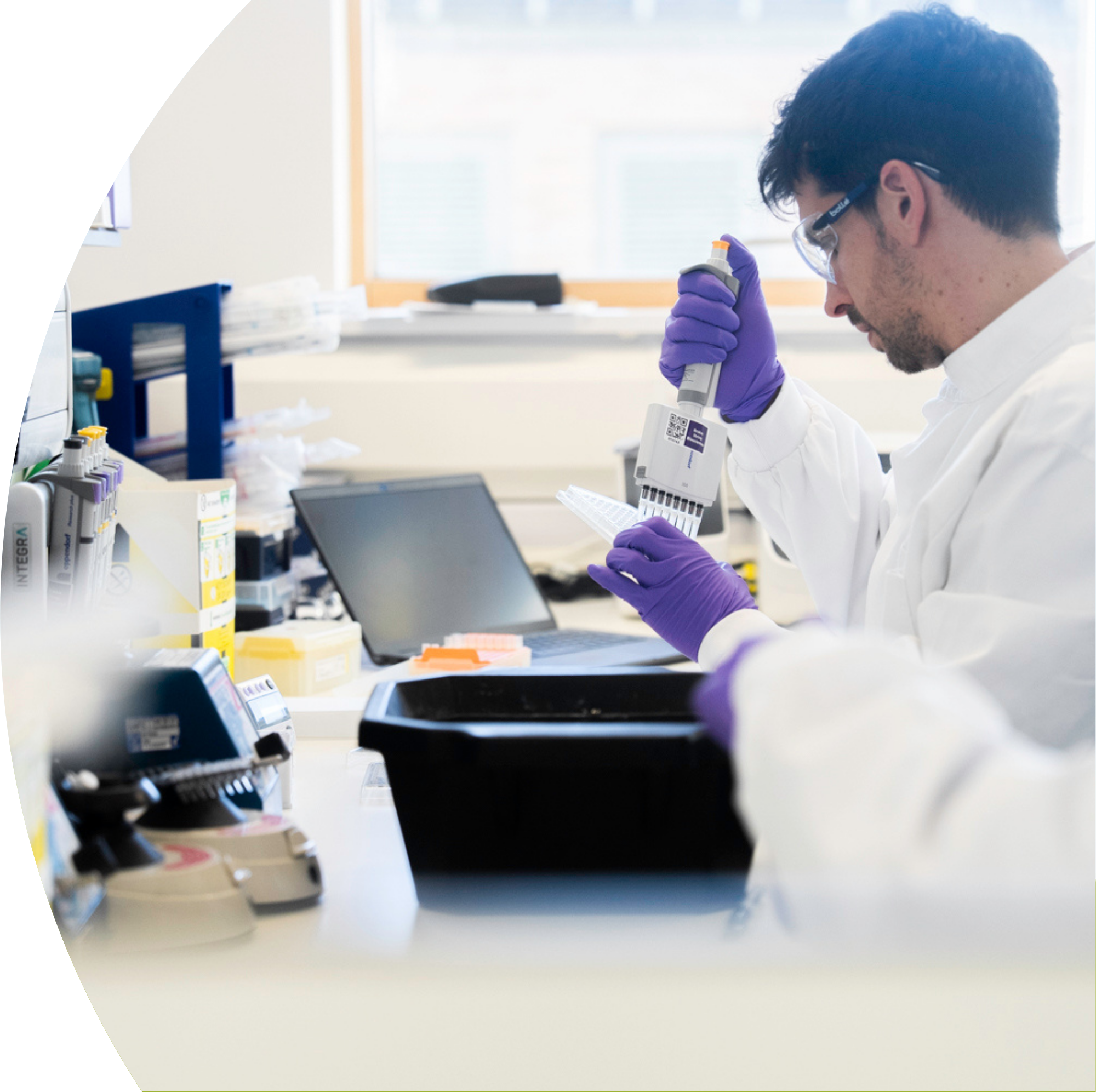 A male scientist wearing a white lab coat, safety goggles, and purple gloves works with a pipette in a laboratory.