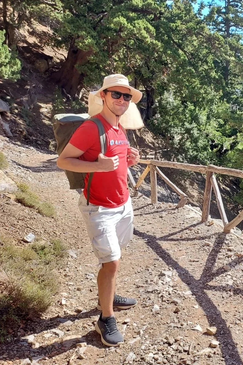 A man hiking on a dirt trail in a forested area, wearing sunglasses, a wide-brimmed hat, a red shirt, beige shorts, and carrying a backpack.
