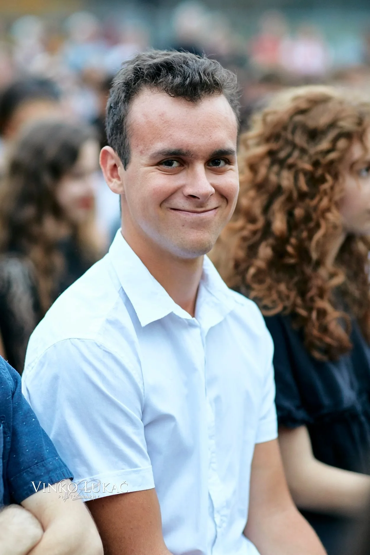 A young man wearing a white shirt sitting among a crowd, smiling at the camera, with other people in the background.