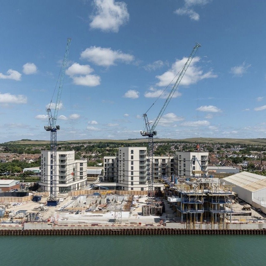 External Mastic. London construction site view with two tall cranes, partially built multi-story buildings, and a body of water in the foreground under a partly cloudy sky.