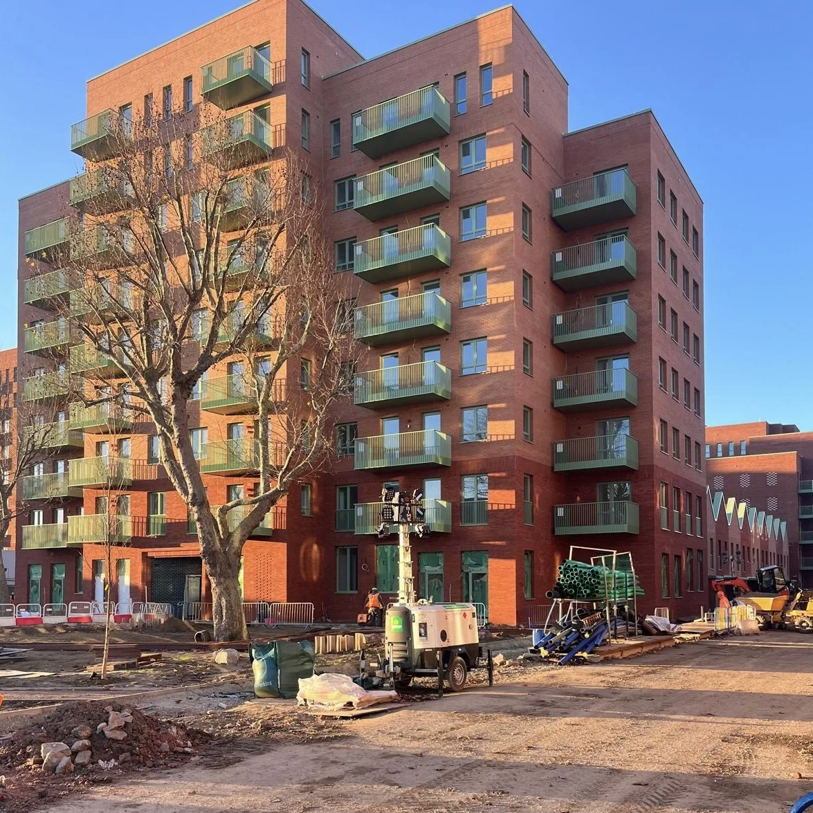 External Window and door mastic work. Red brick apartment building under construction with green balconies, surrounding construction site with equipment, a leafless tree, and a blue sky.