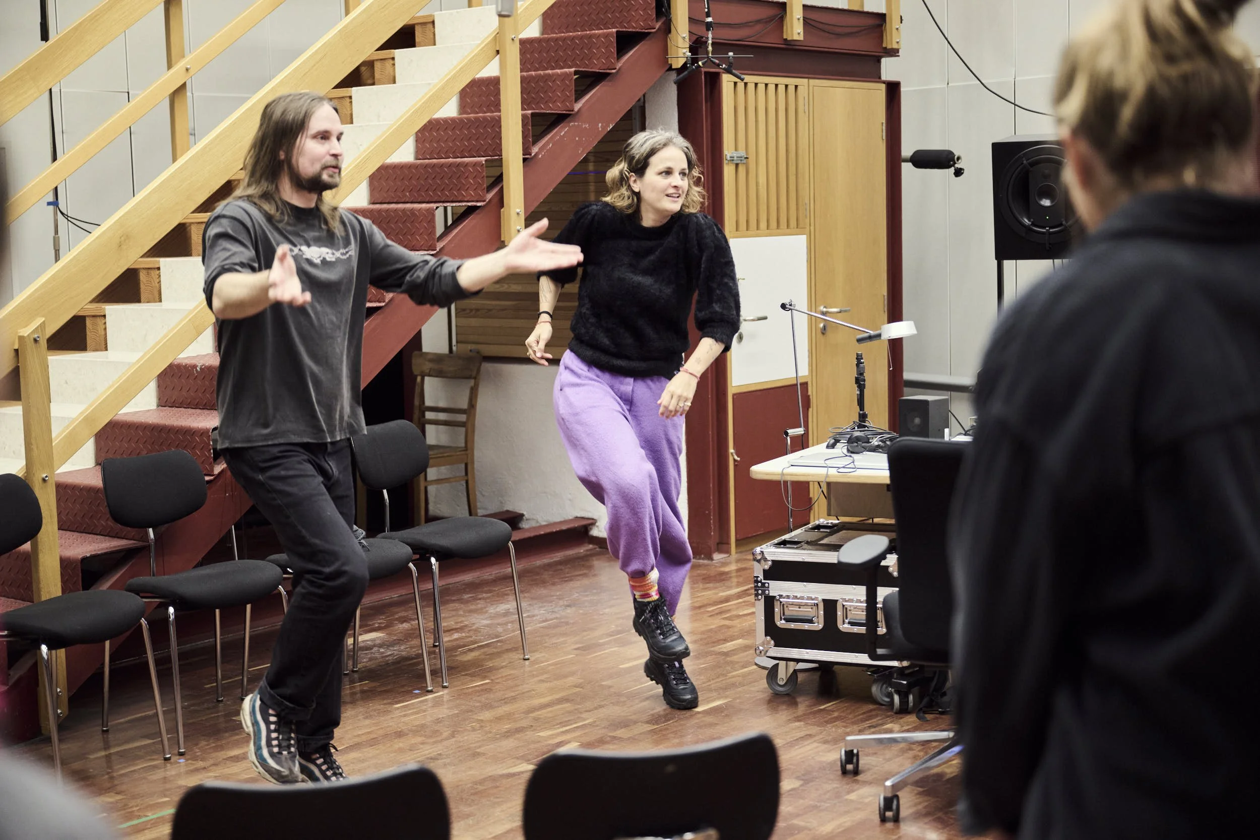 Two women are dancing and a man is gesturing in an indoor rehearsal or studio space with a wooden staircase, chairs, and sound equipment.