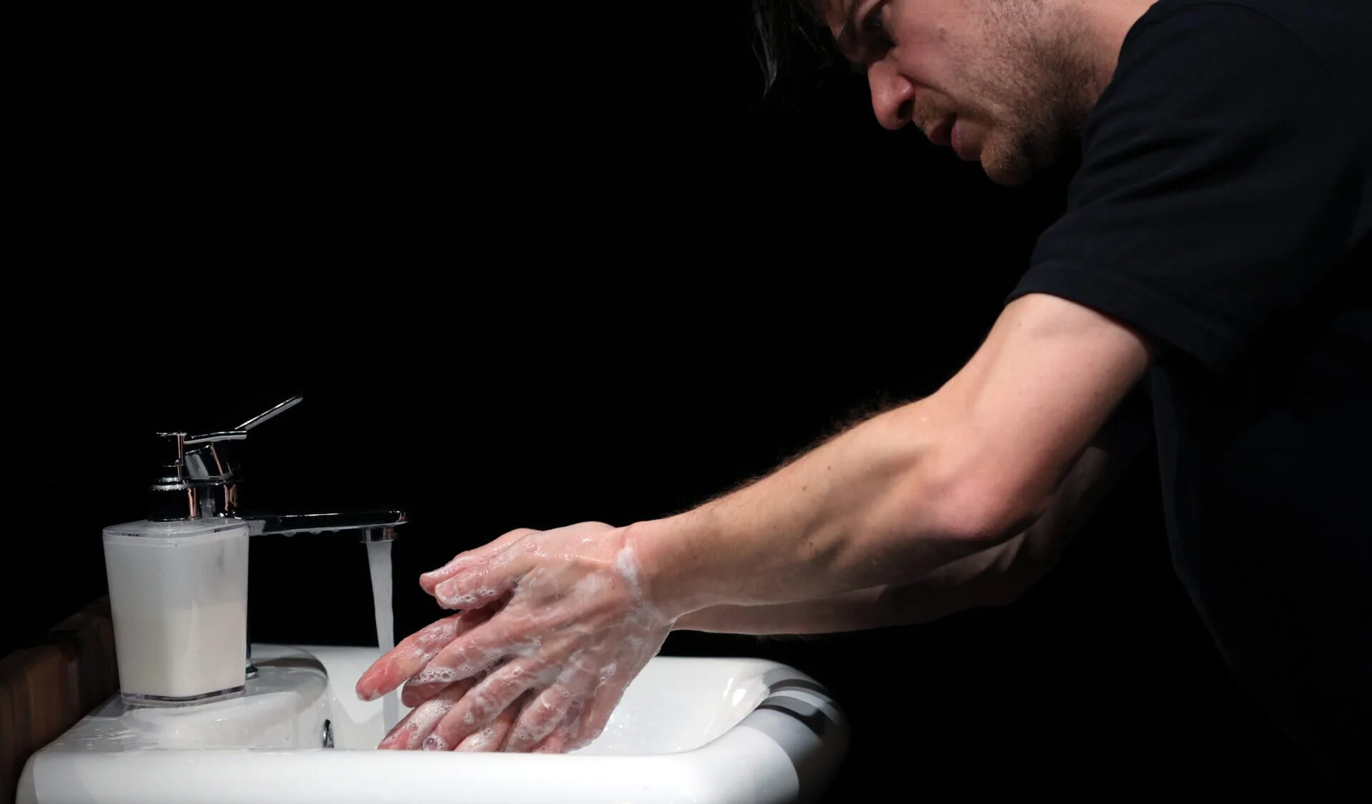 A man washing his hands at a sink with soap lather, with a black background.