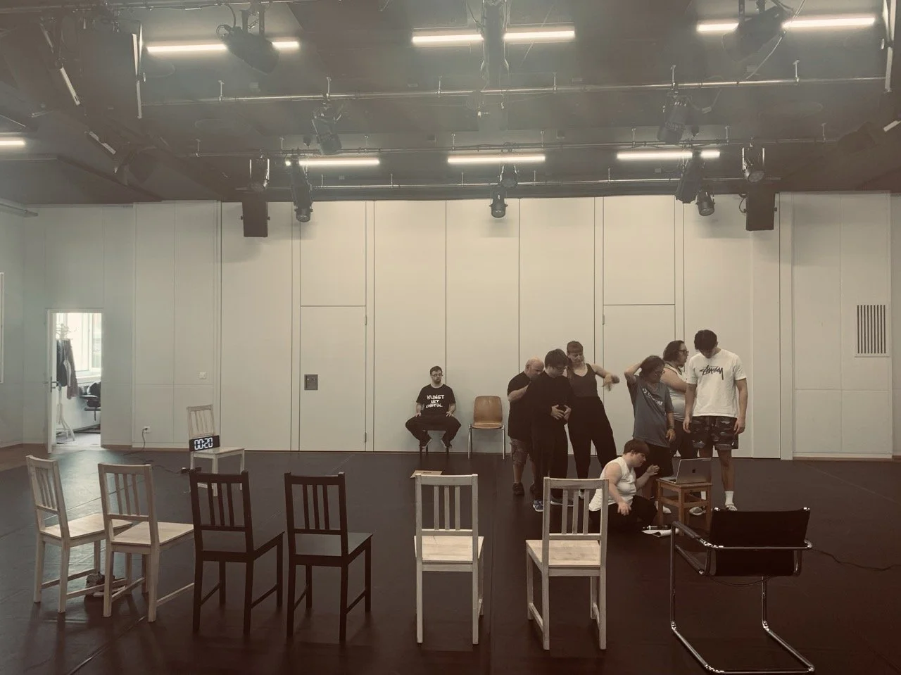 A group of people in a large room with a dark floor, some standing and some sitting, around a laptop on a small table. Seven chairs are arranged in the foreground, and a digital clock shows 4:20.