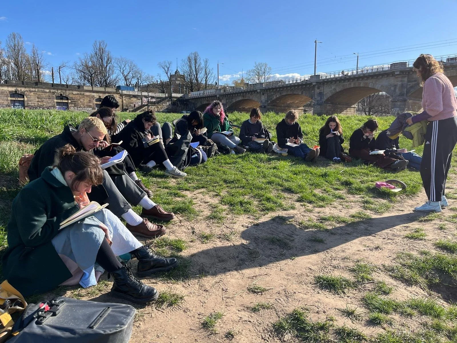 A group of students sitting on the grass, reading books outside on a sunny day near a bridge.