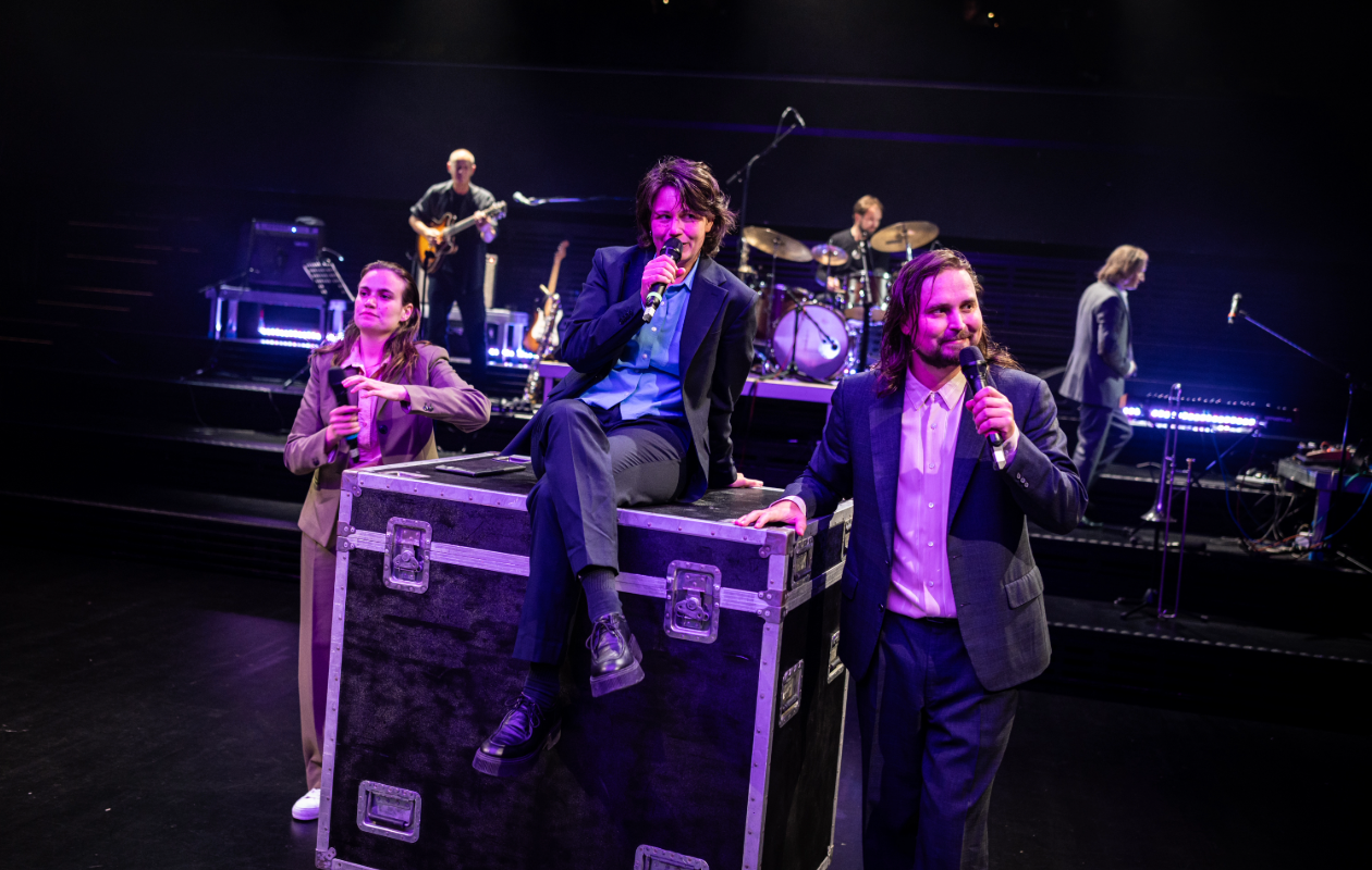 People with microphones, a woman sitting on a box, a man standing beside her, and a band playing instruments on stage with drums and guitars.