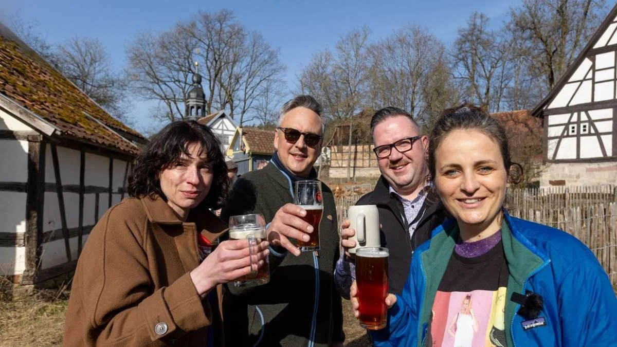 Four people outdoors in front of traditional half-timbered houses enjoying drinks on a sunny day.