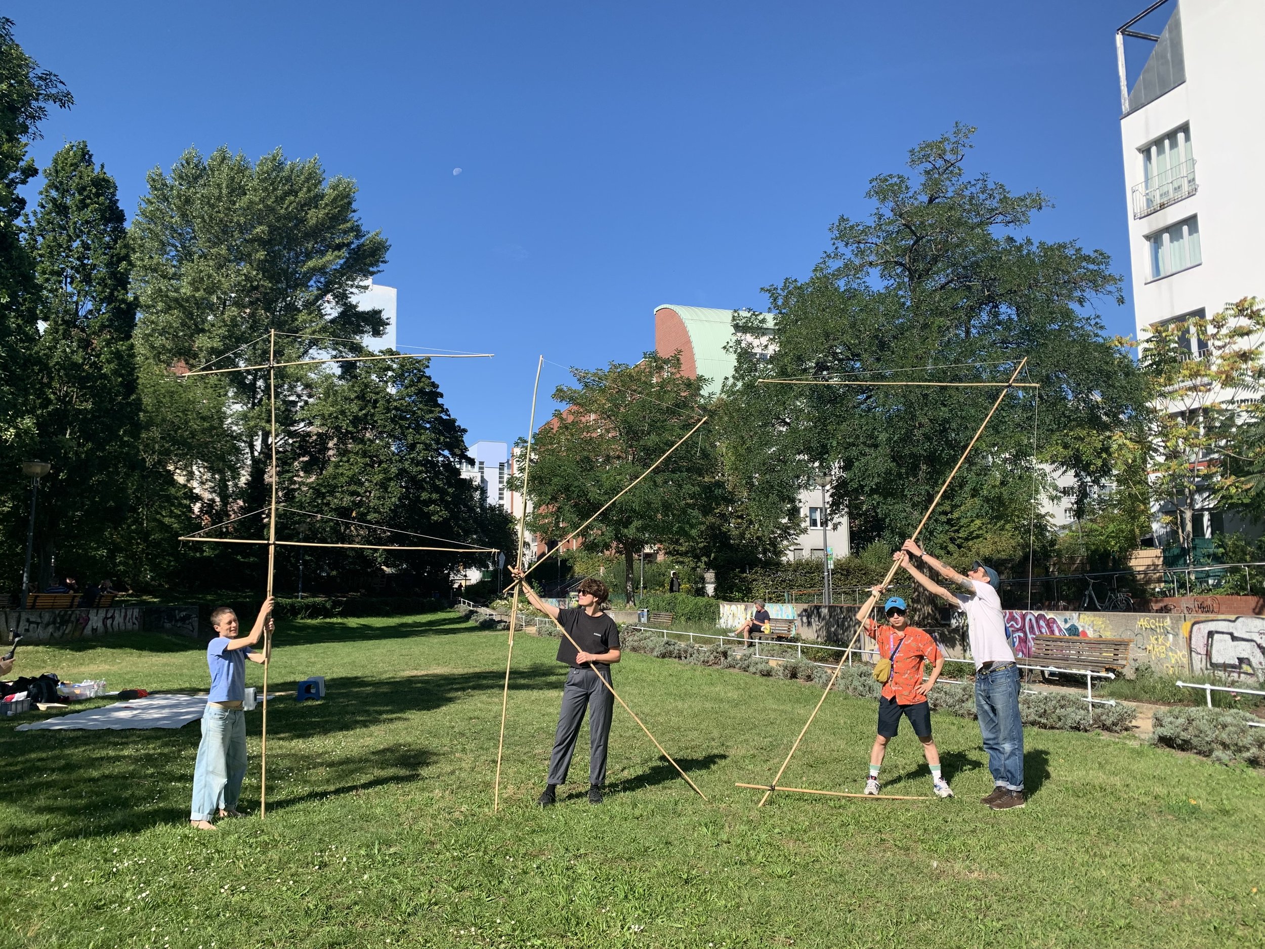 Four people in a park setting using long poles to support a large, square-shaped structure made of thinner poles, against a background of trees, buildings, and a bright blue sky.